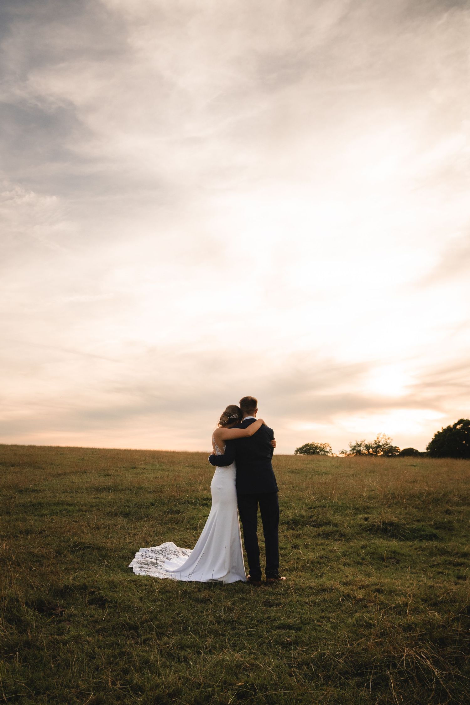 A couple embraces in a romantic sunset silhouette on a grassy hilltop with their wedding attire flowing in the breeze.