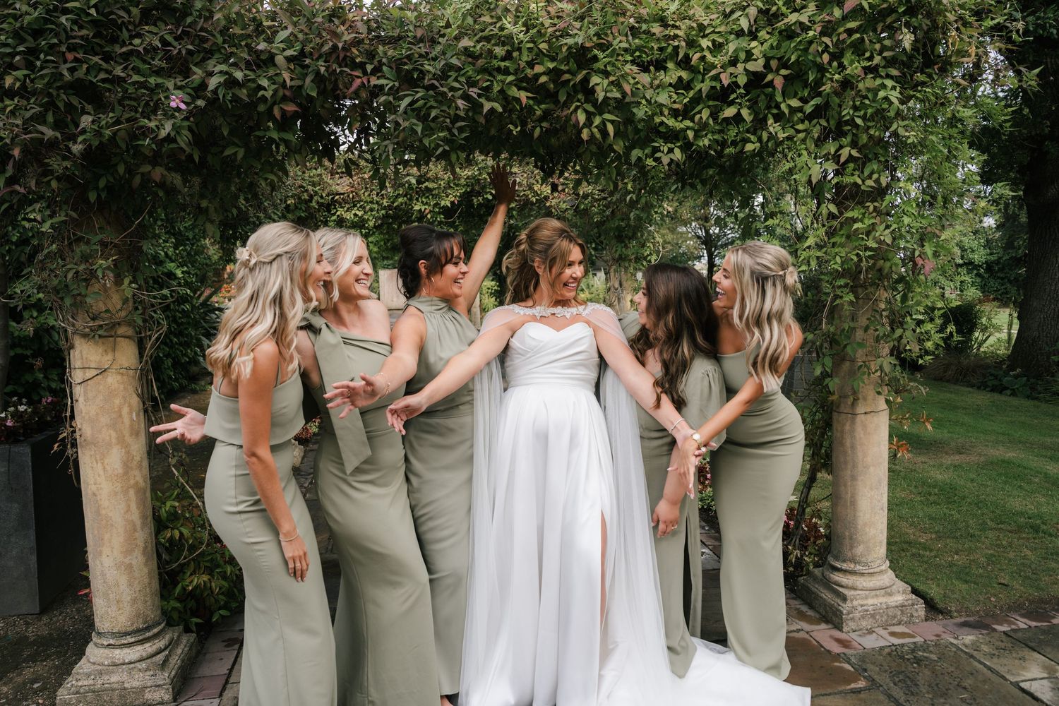 Wedding party in sage green dresses poses together against lush garden setting.