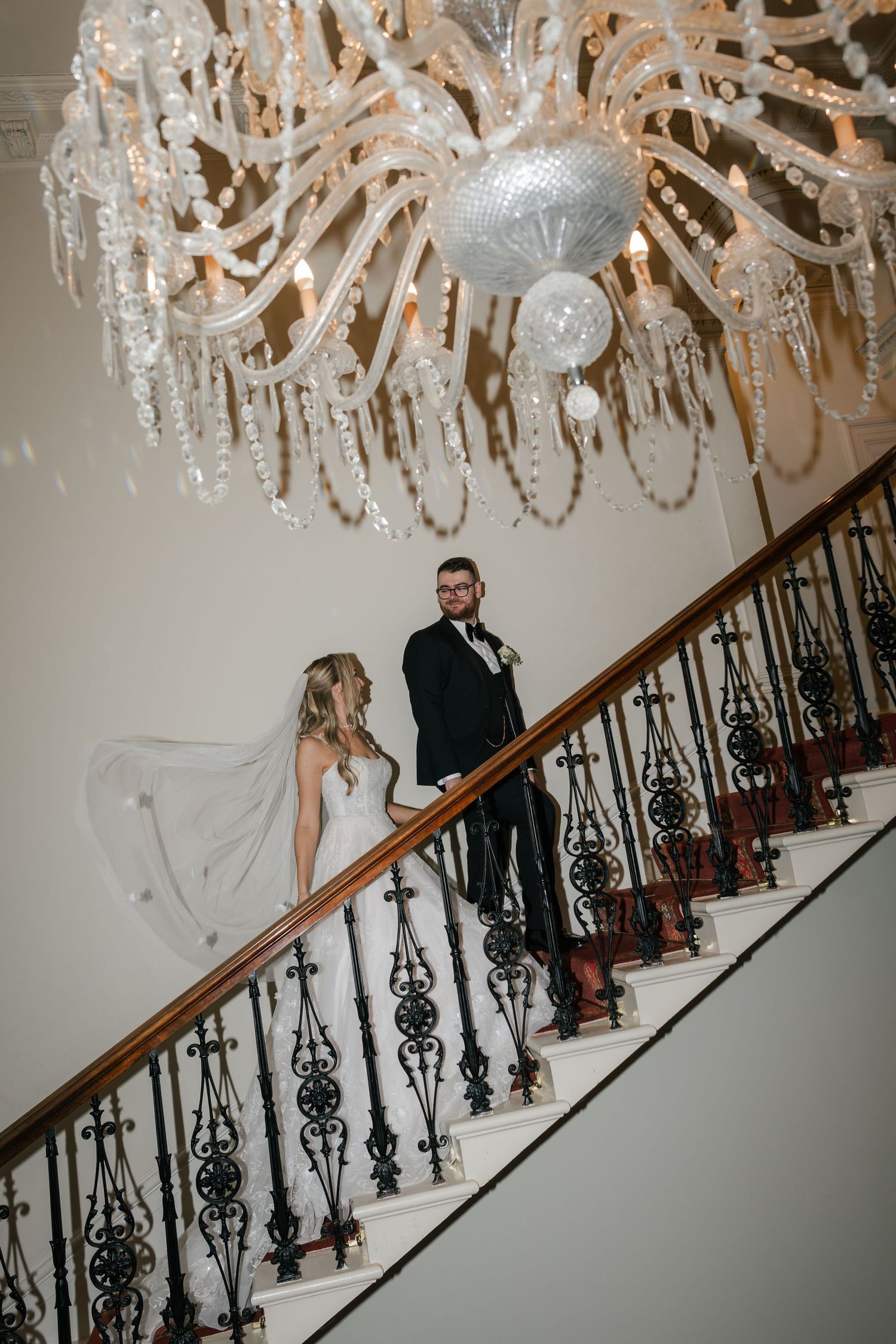 A bride and groom descend an elegant staircase under a crystal chandelier in a romantic wedding moment.