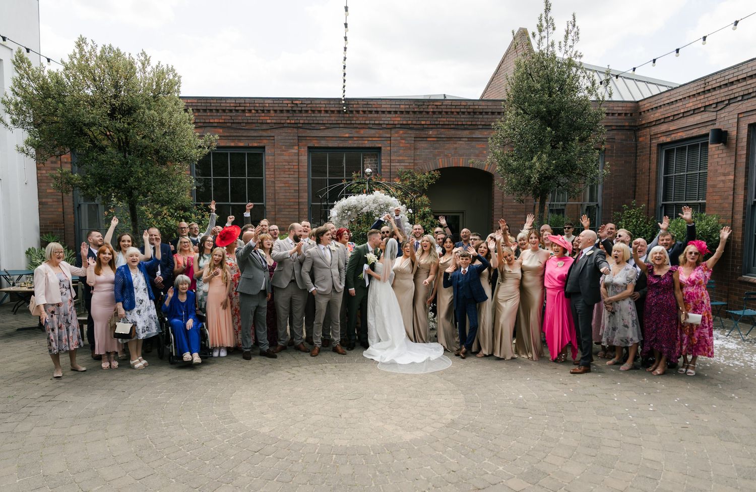 A panoramic wedding group photo showing a large gathering of guests and wedding party outside a brick building.