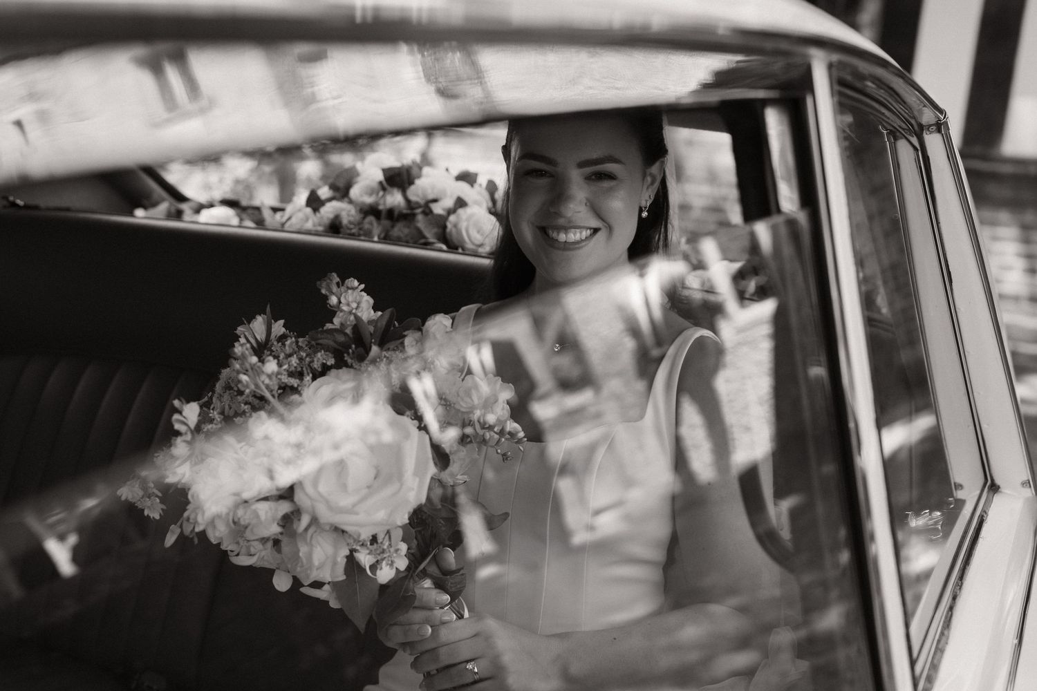Smiling bride holding a floral bouquet inside a vintage car on her wedding day, black and white photo.