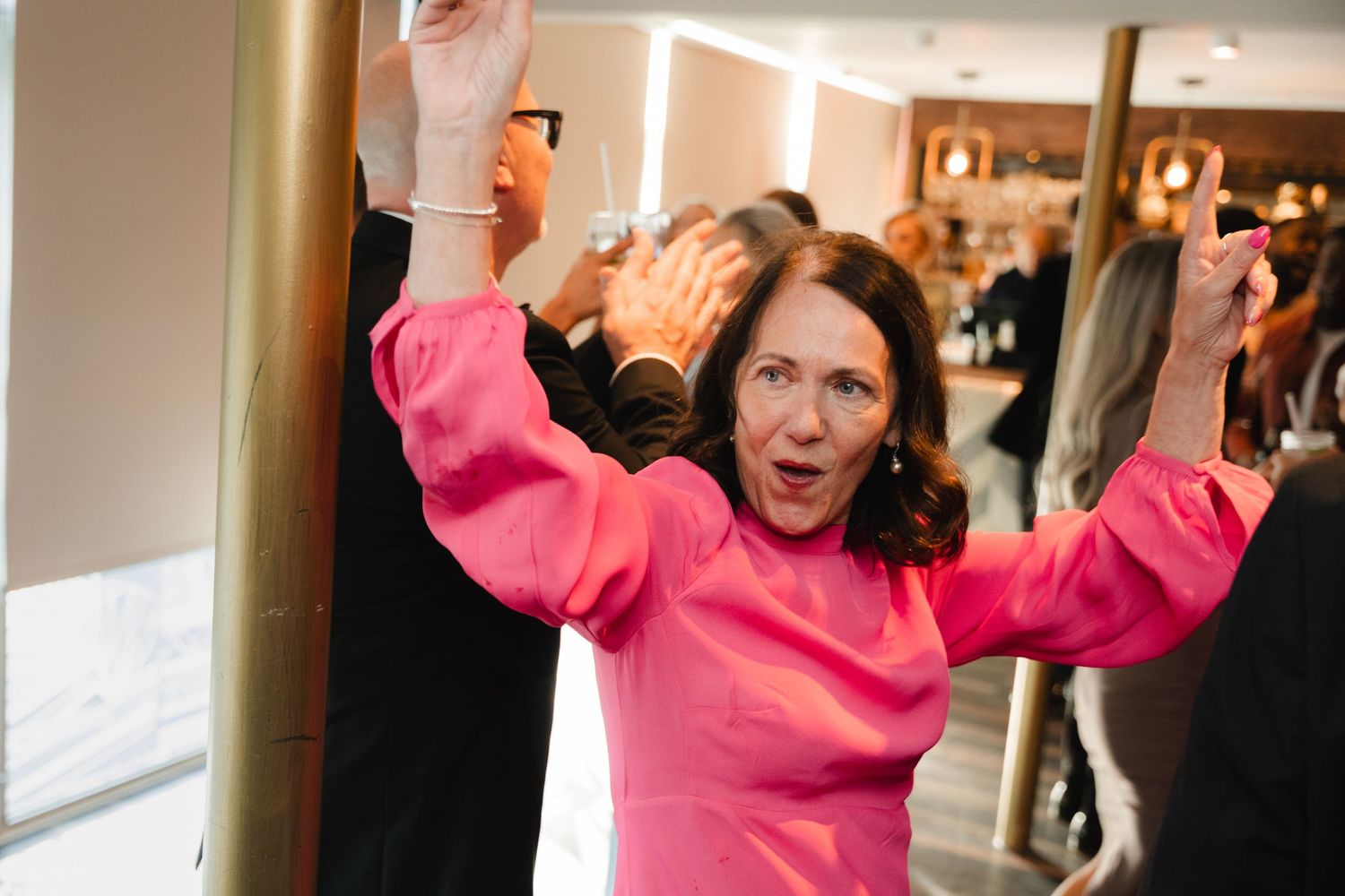 A person in a bright pink sweater raises their arms while dancing or celebrating in an indoor setting.