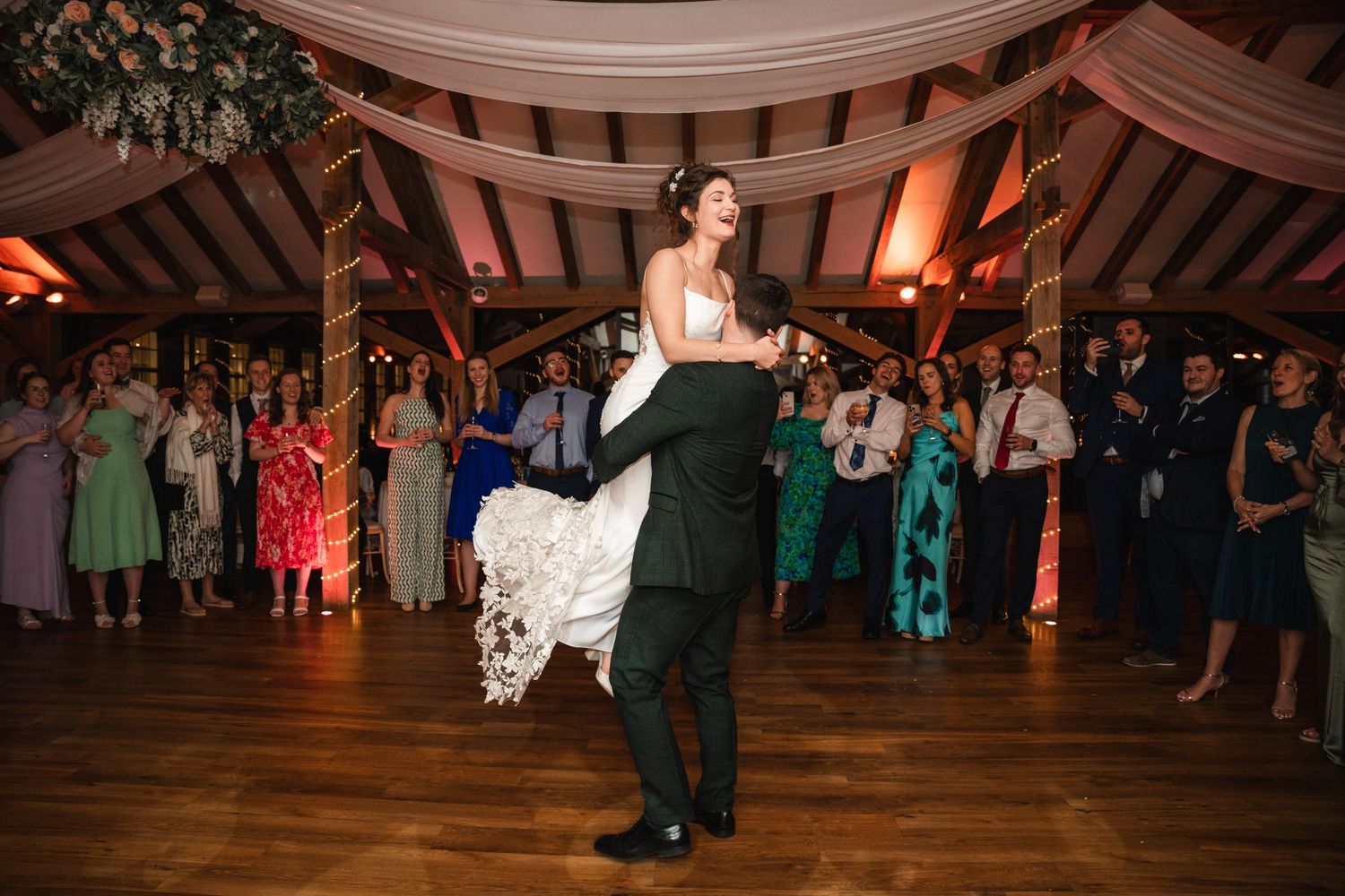 Wedding guests dance and celebrate on an elegant dance floor under warm lighting and decorative architectural details.