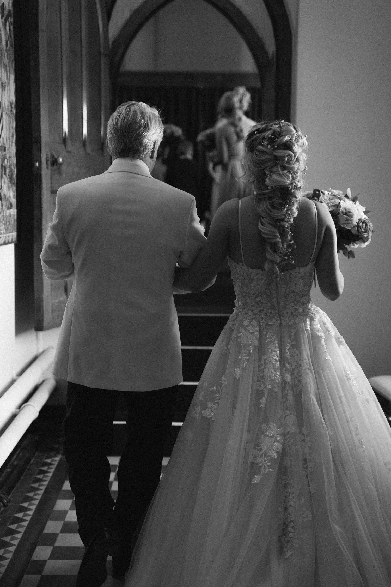 Black and white wedding photo showing a bride in a lace-detailed gown walking up stairs with a companion.