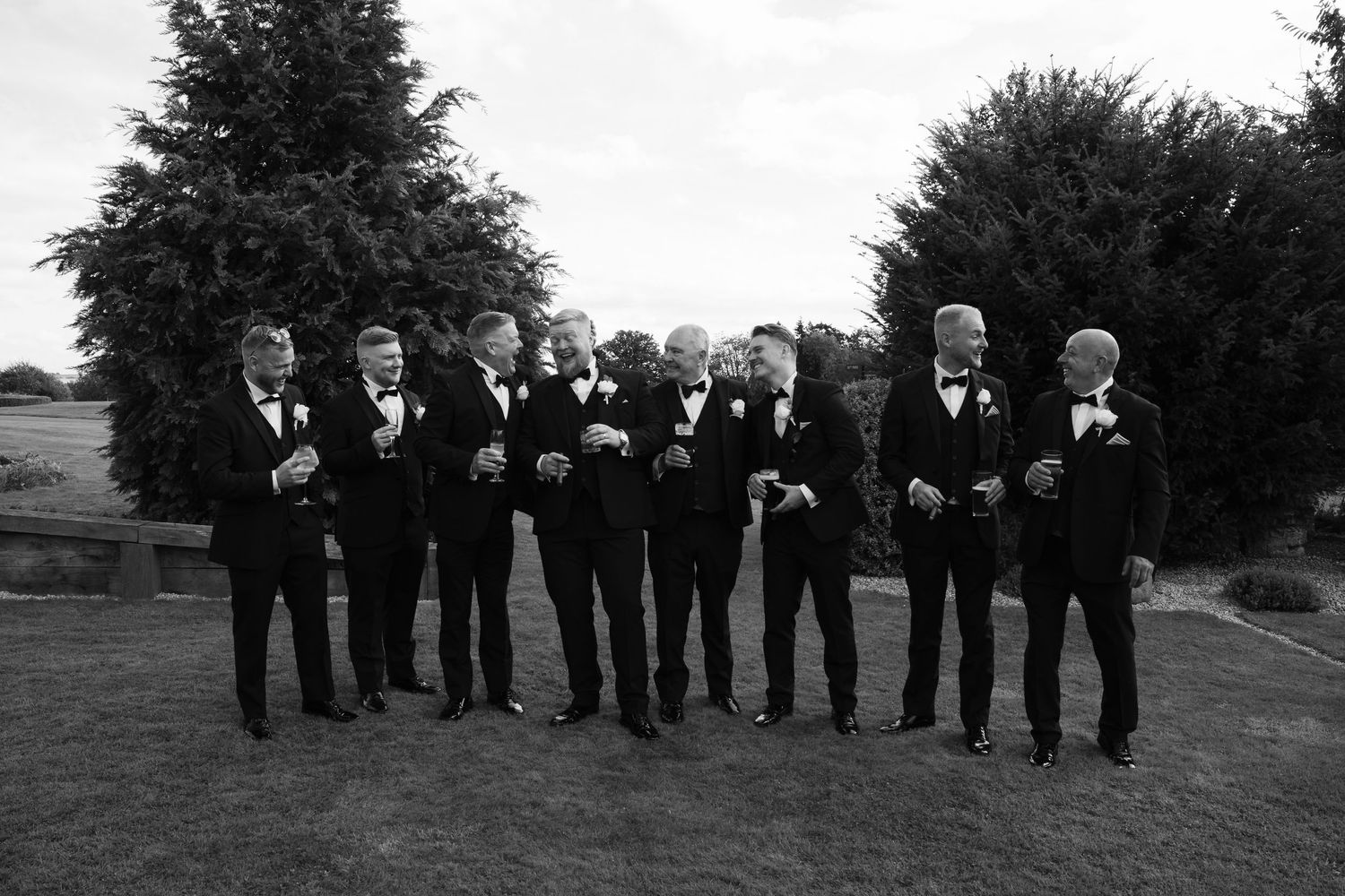 Wedding party in formal black attire stands in line outdoors against trees and greenery.