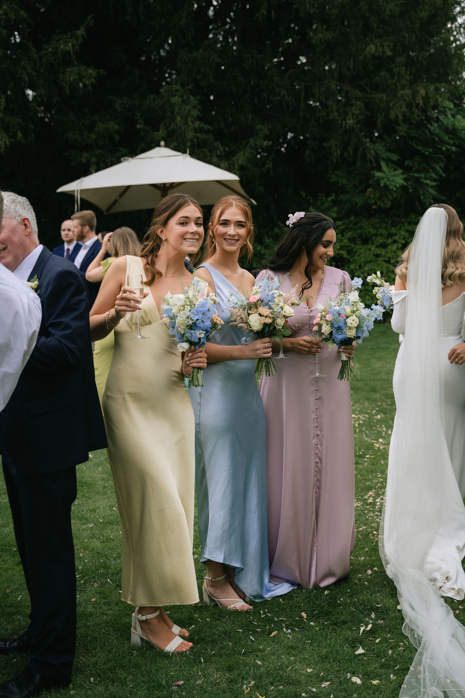 Wedding party stands in a line on grass wearing light blue, beige and white formal attire while holding floral bouquets.