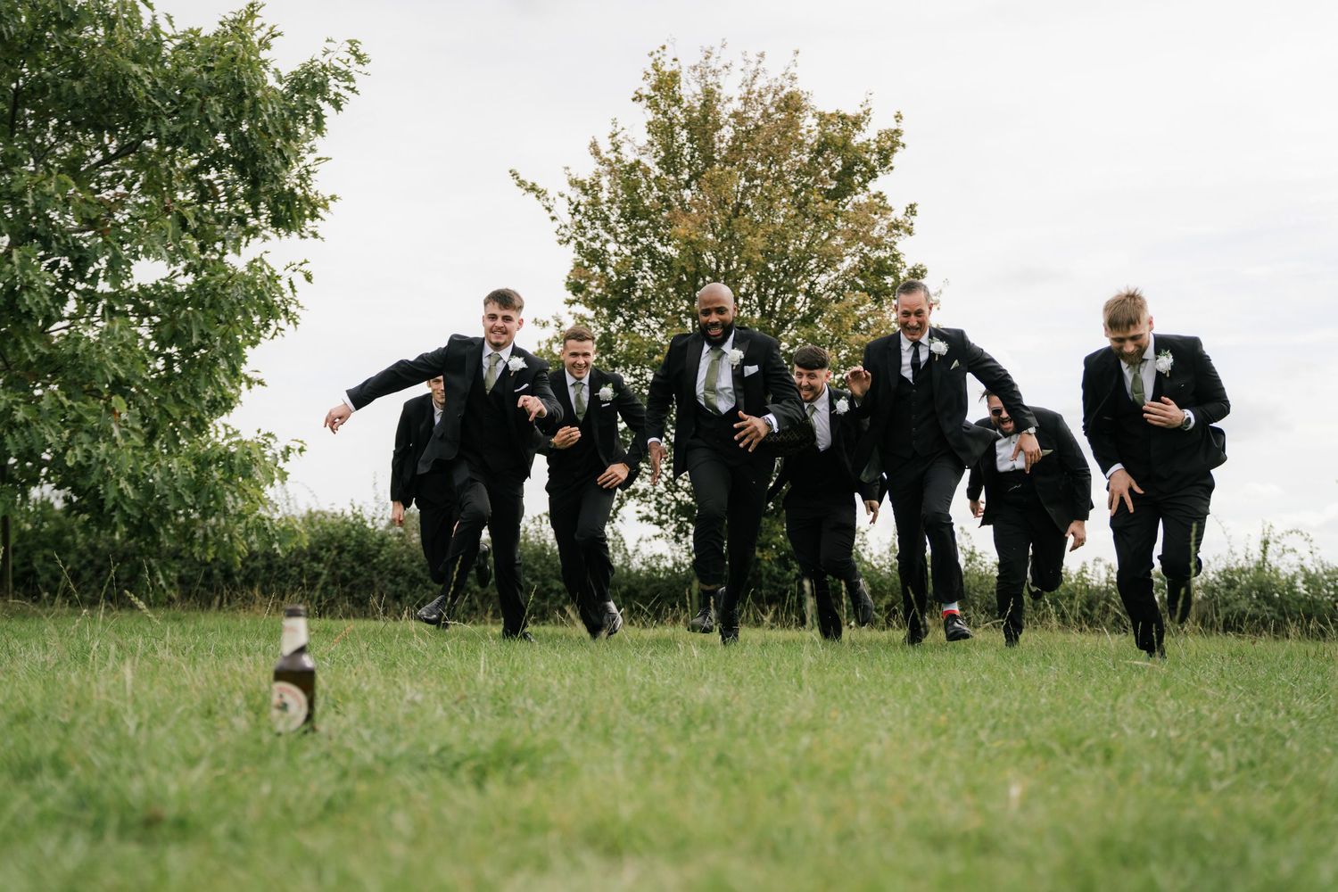 Groomsmen in black suits run across a grassy field during wedding festivities.