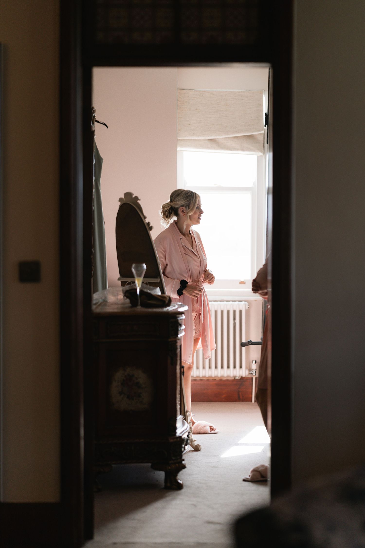 A silhouetted figure stands in a sunlit doorway of a vintage room with antique furniture.