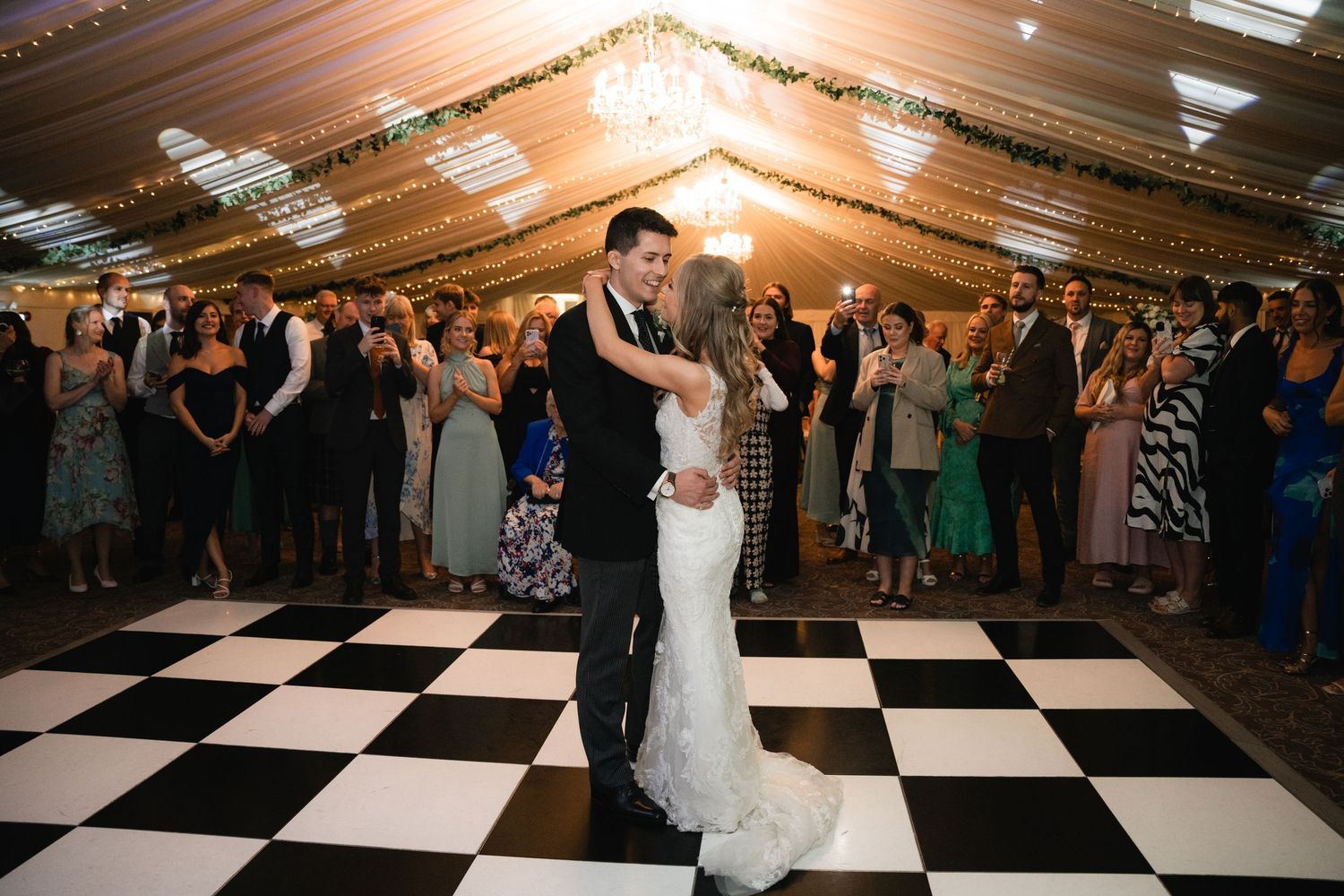 A couple shares their first dance on a black and white checkered floor under a tent with string lights and wedding guests.