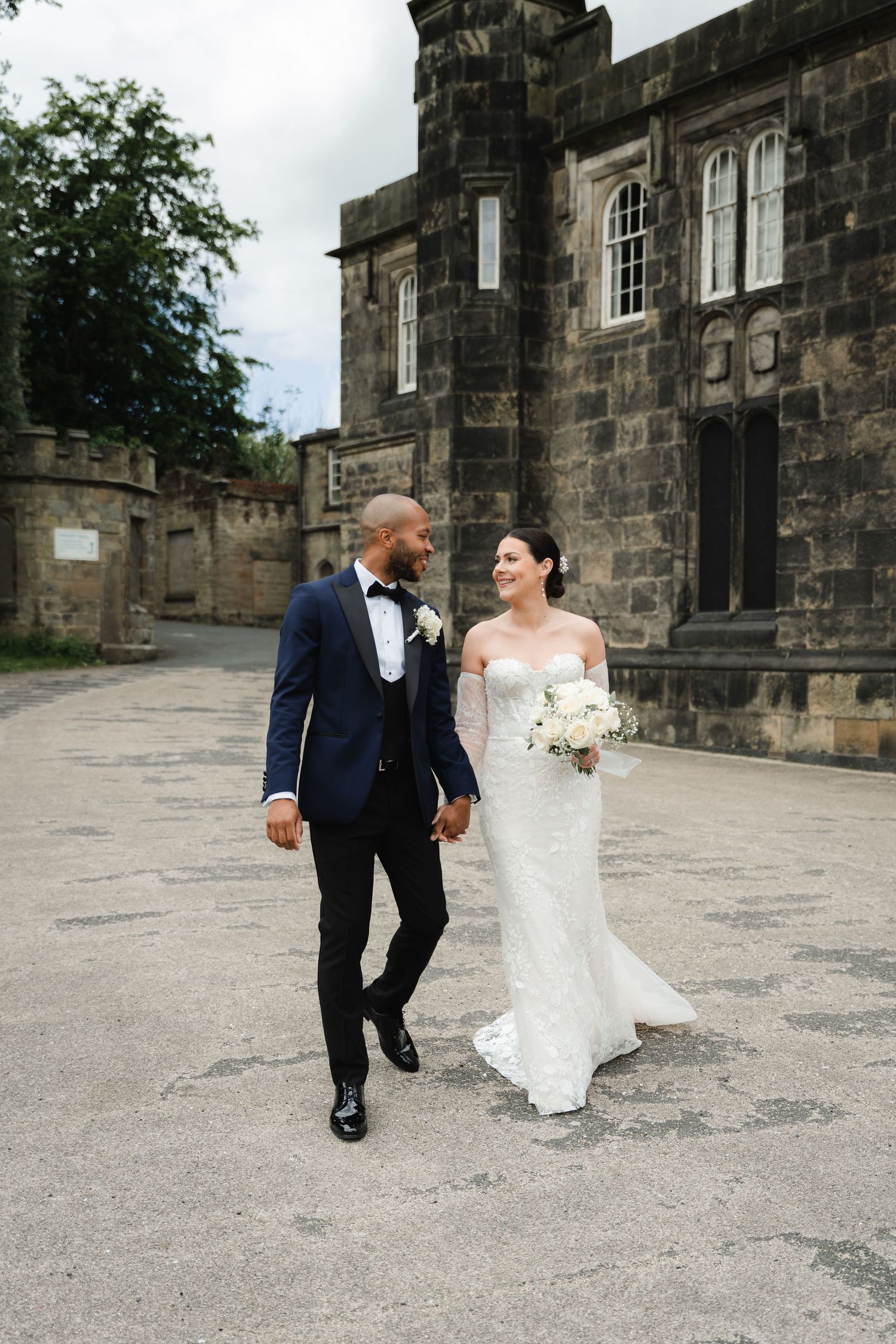 A couple in wedding attire walks and dances together on the stone courtyard of a historic building.