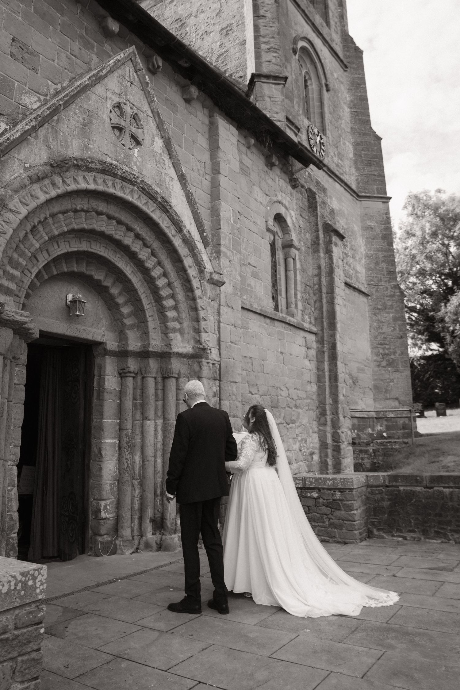 A couple stands in front of a historic stone church entrance with ornate Romanesque archways in black and white.