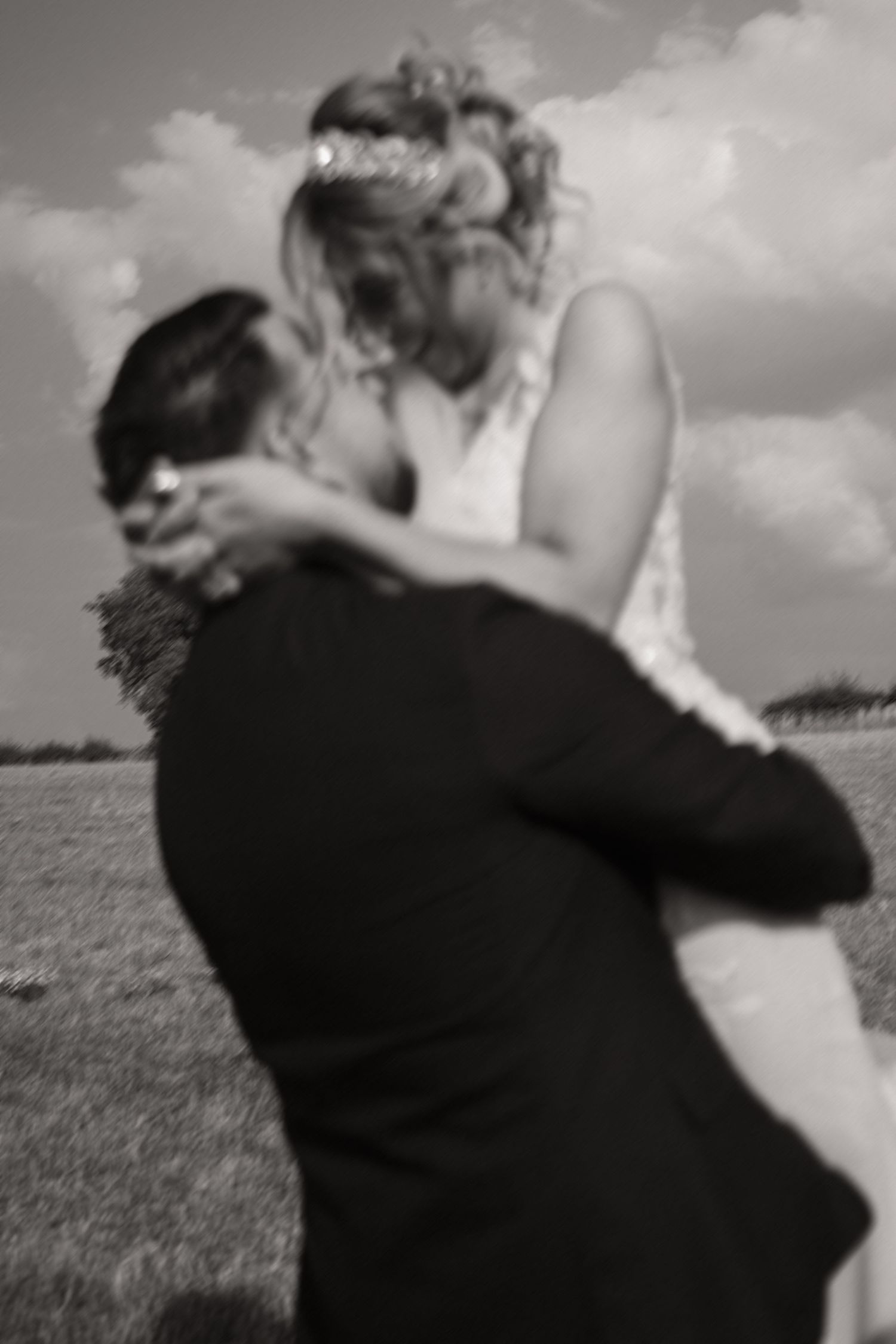 A romantic black and white wedding photo shows a couple embracing in an outdoor field under cloudy skies.