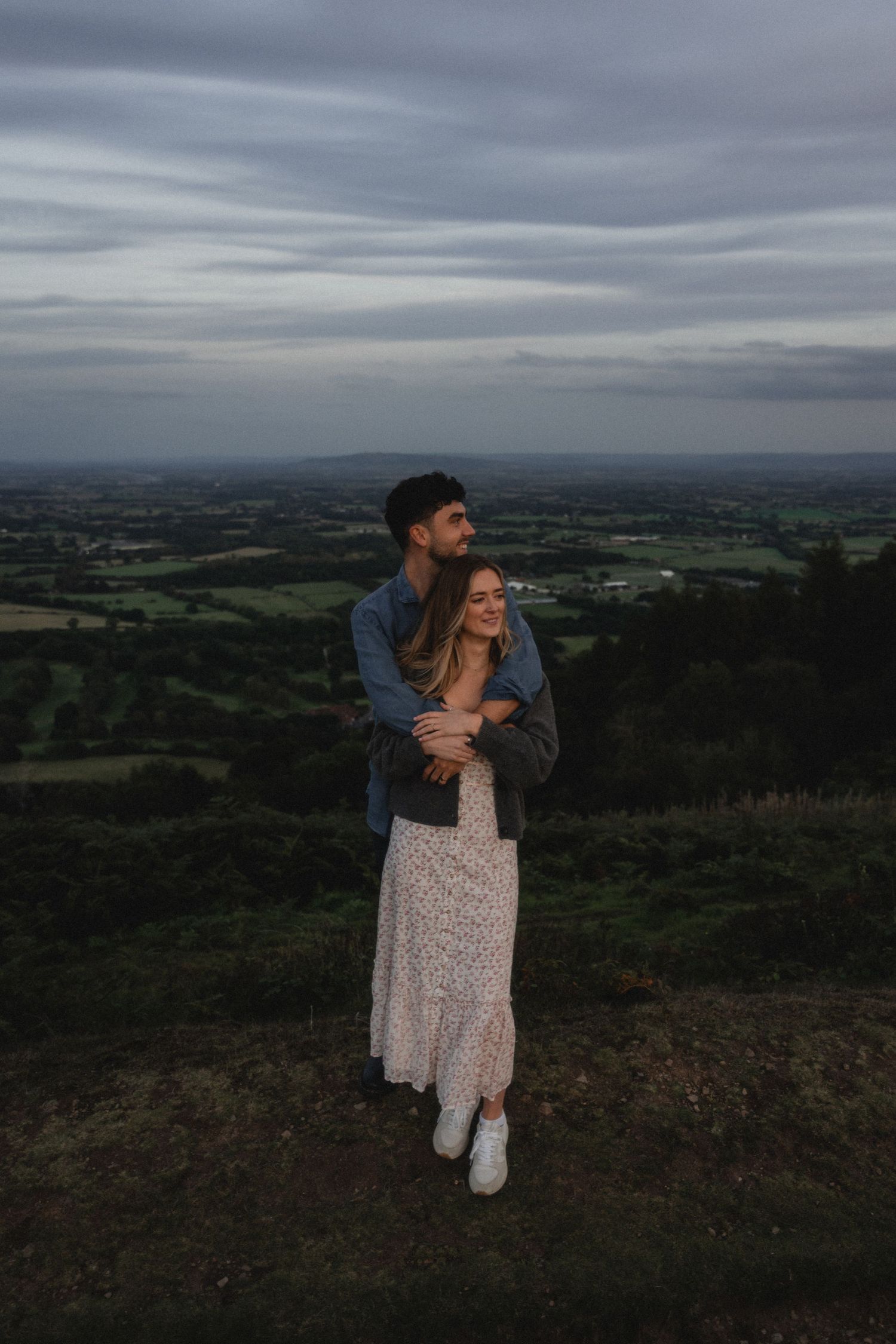 A couple embraces while enjoying panoramic views of rolling hills and farmland at dusk.