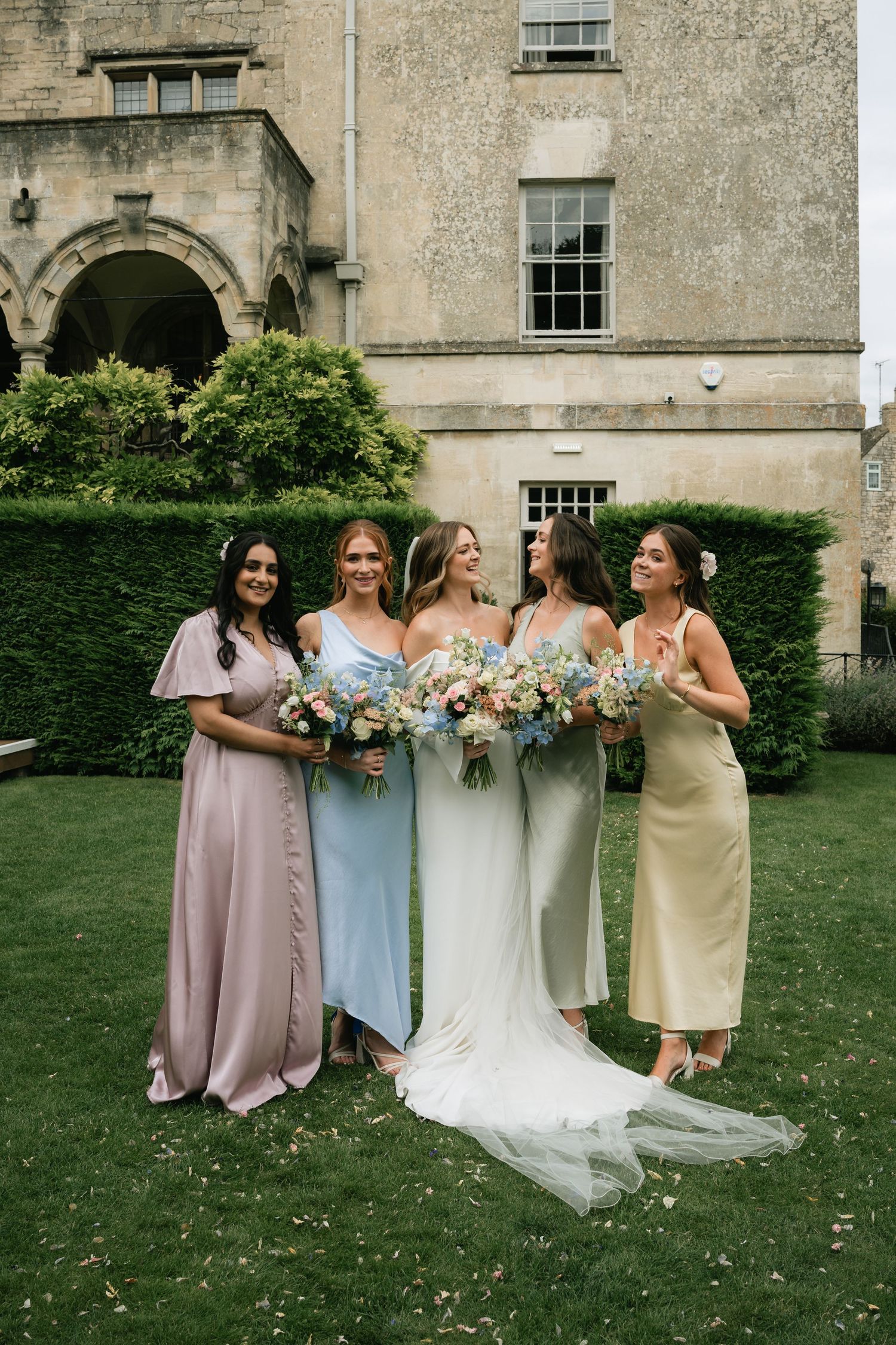 A bridal party stands on a manicured lawn in front of a historic stone building with pastel dresses and floral bouquets.