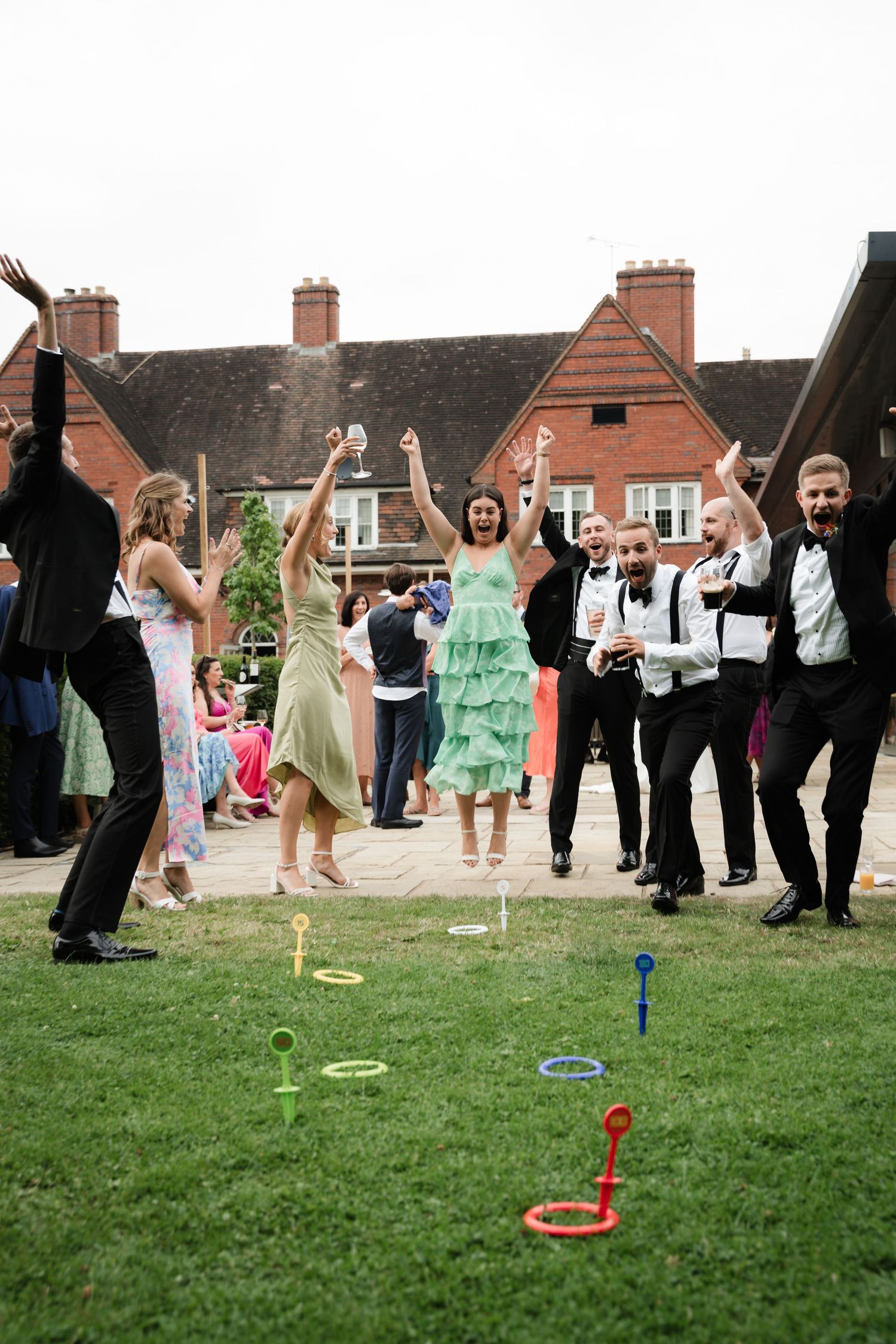 Wedding guests play lawn games and celebrate outdoors on a grassy area near brick houses.