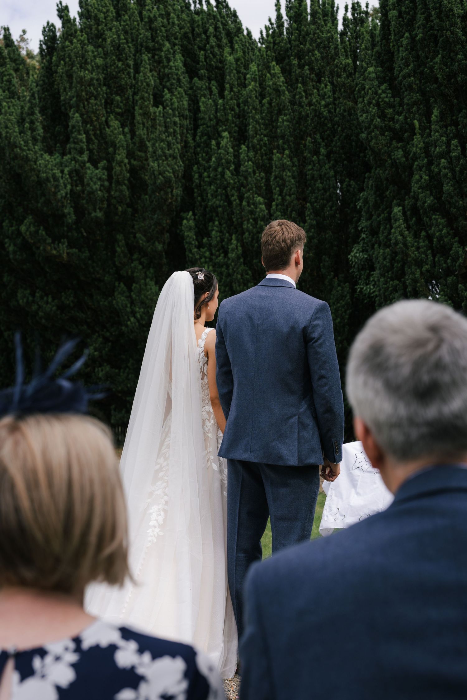 Wedding guests watch as a couple stands together at an outdoor ceremony surrounded by tall evergreen trees.