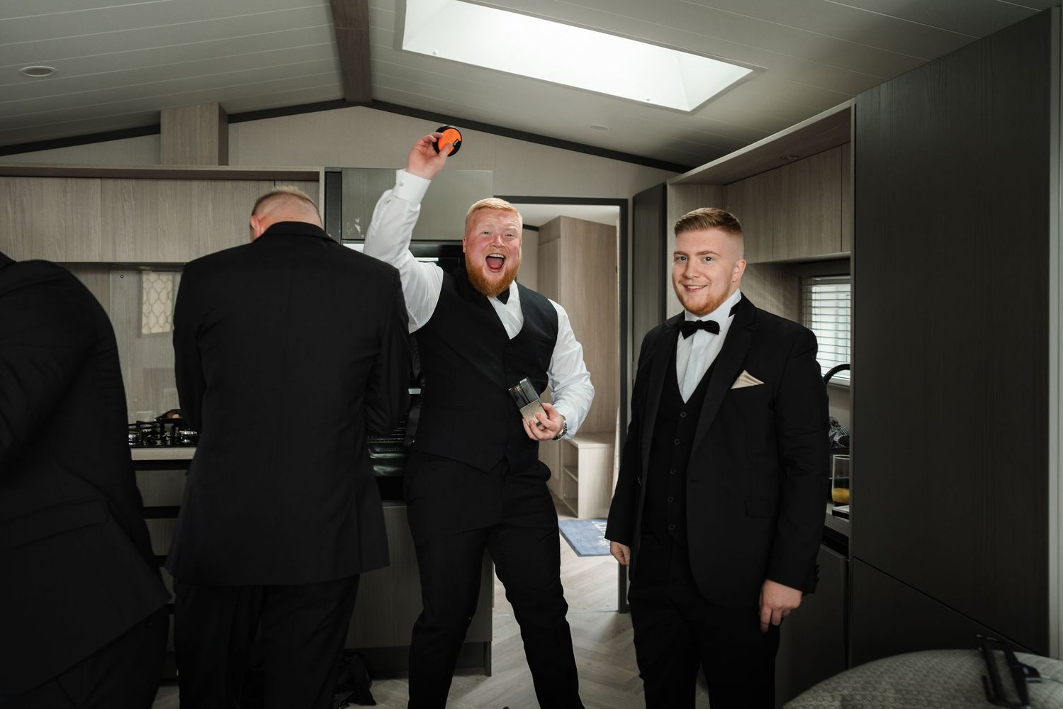 Men in formal black tuxedos gather in modern room with skylight.