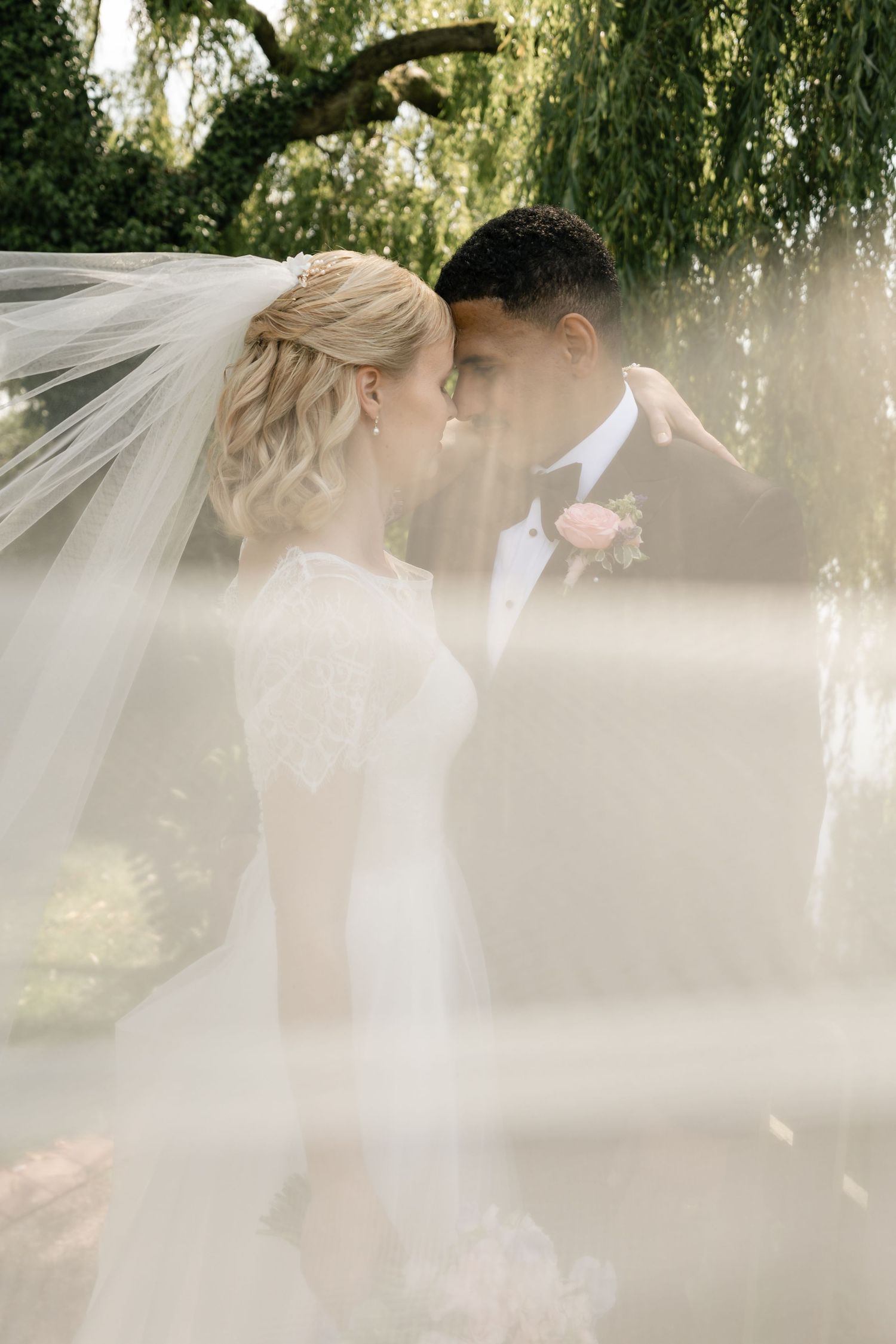 Ethereal wedding portrait under a weeping willow tree with flowing veil in soft natural light.