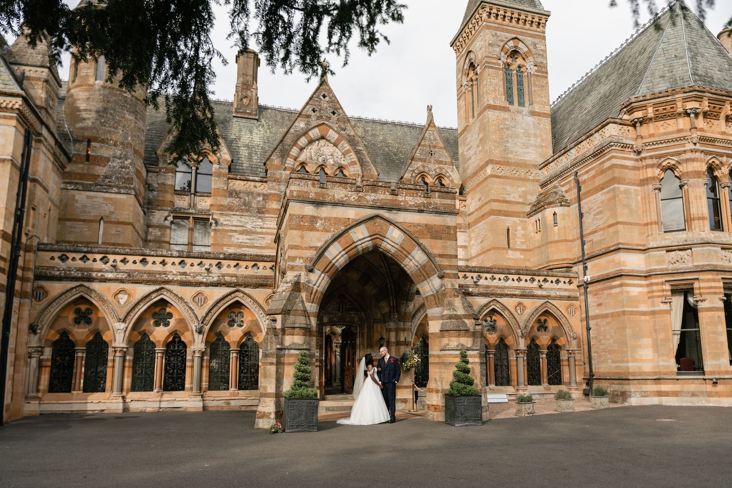 A couple stands in the ornate Gothic archway entrance of a historic sandstone church with Victorian architectural details.