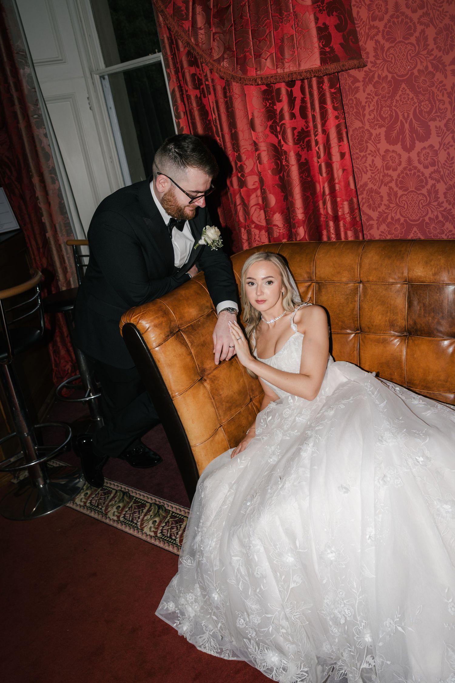 A couple poses on a vintage leather couch against red damask wallpaper in a series of wedding photos.