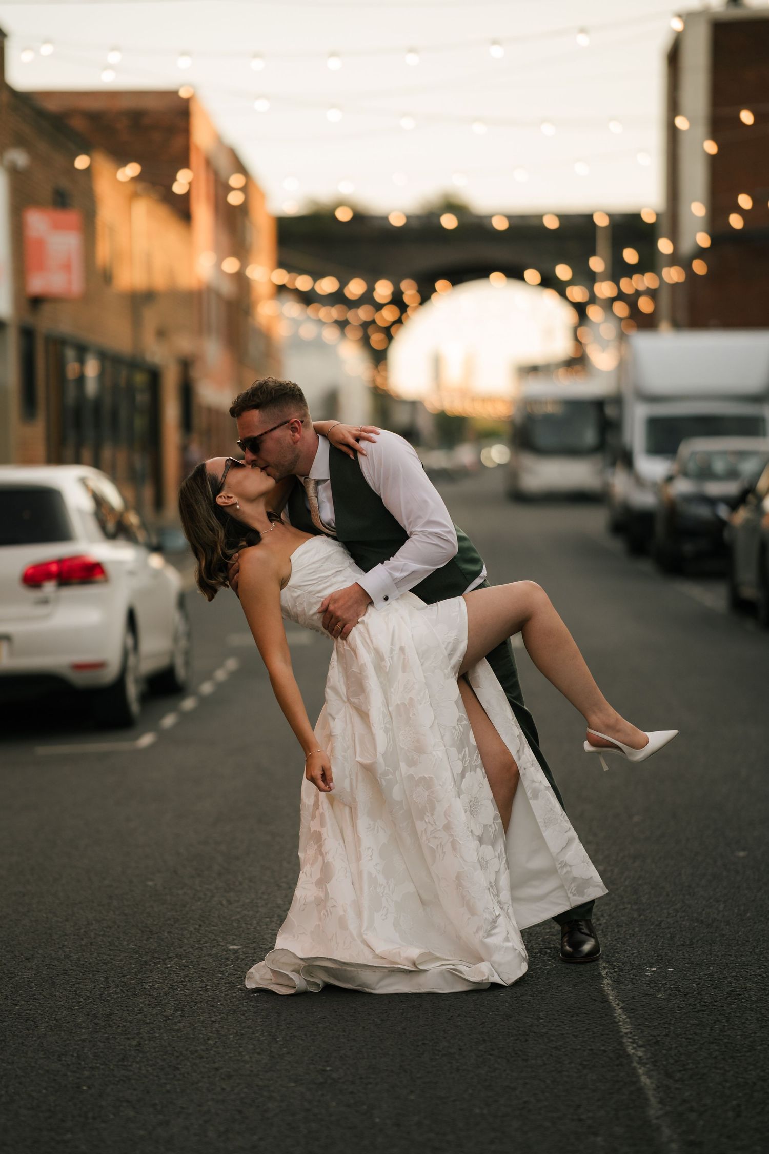 A romantic dip pose between a couple in wedding attire on a city street with string lights in the background at dusk.