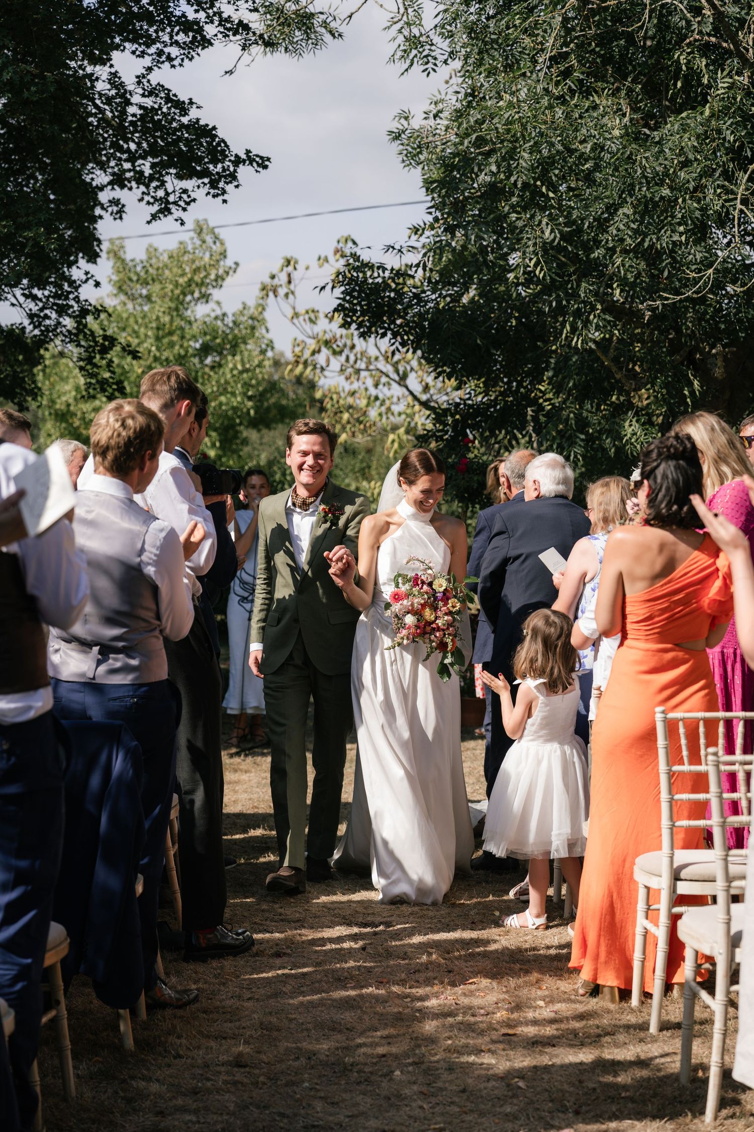 Wedding guests gather outdoors under trees at a rustic ceremony with white chairs visible in the background.