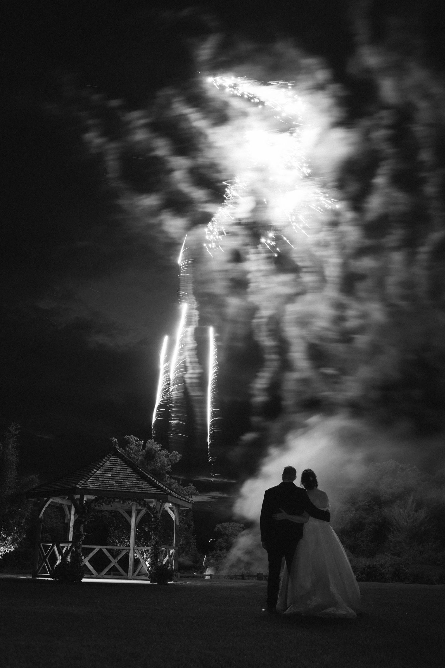 Silhouettes of people watching a dramatic fireworks display over a gazebo structure in black and white.