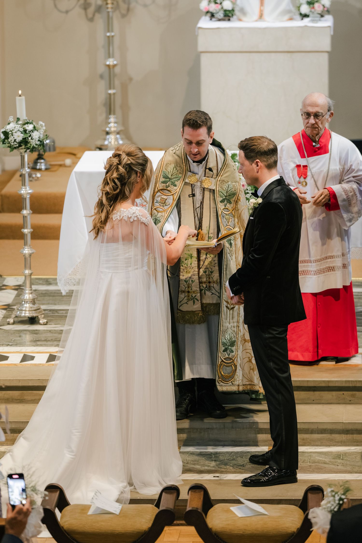 Traditional Catholic wedding ceremony taking place at church altar with priest in ornate vestments and couple exchanging vows.