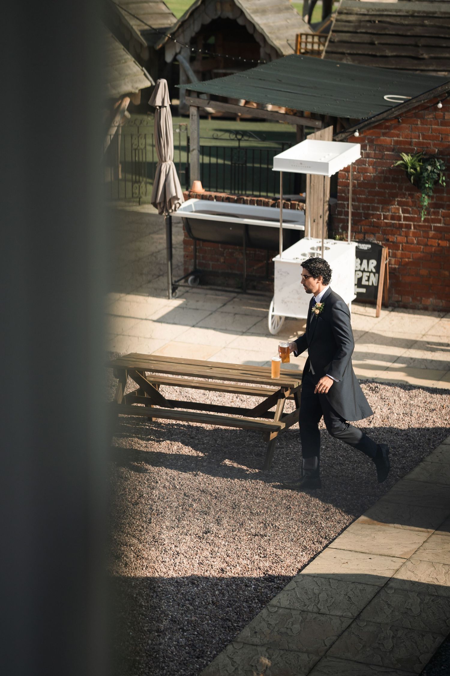 A person in dark formal attire walks through a courtyard with brick walls and outdoor furniture on a sunny day.