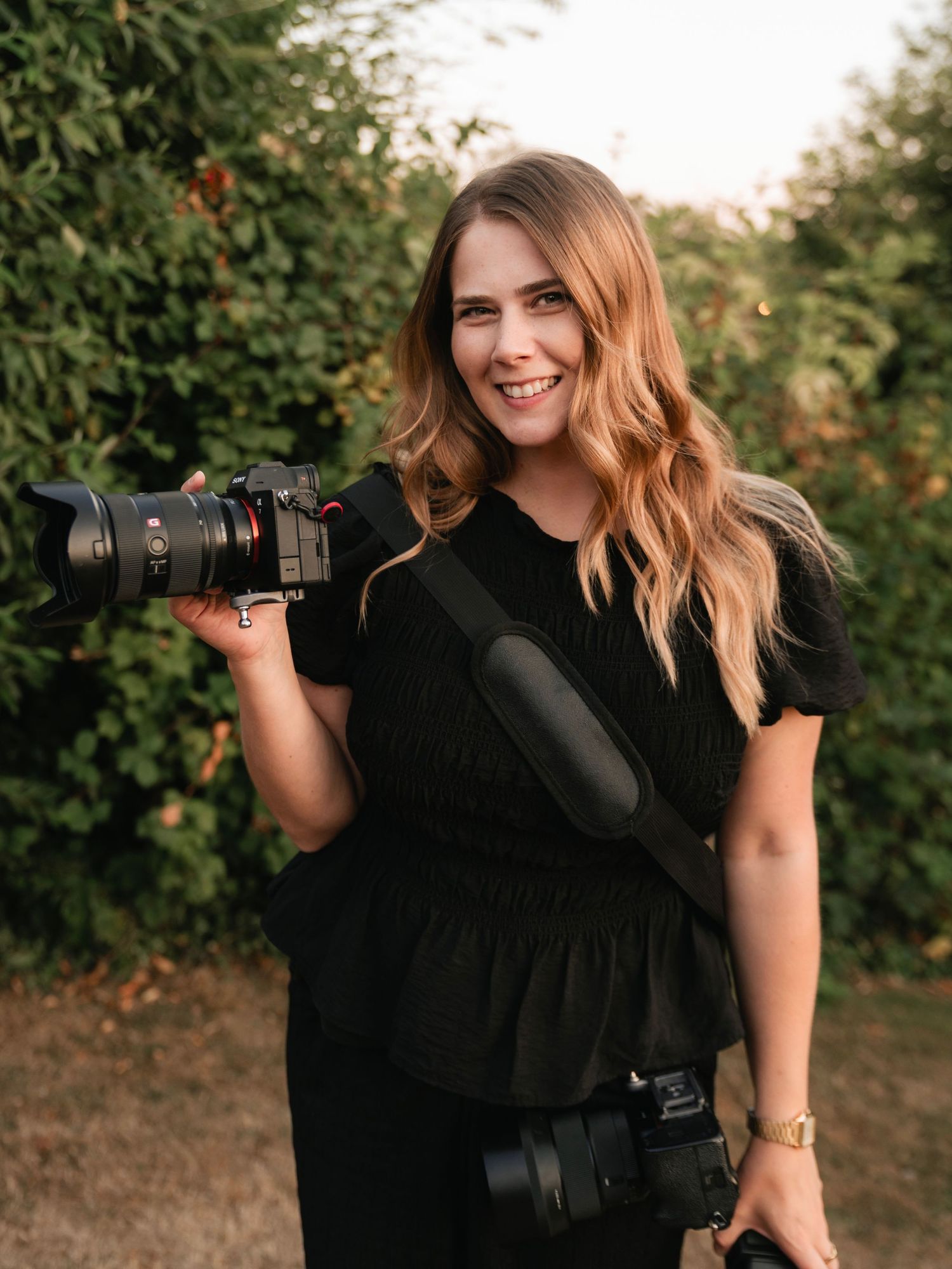 Smiling female photographer holding a professional camera with lens, wearing black outfit outdoors near green trees.