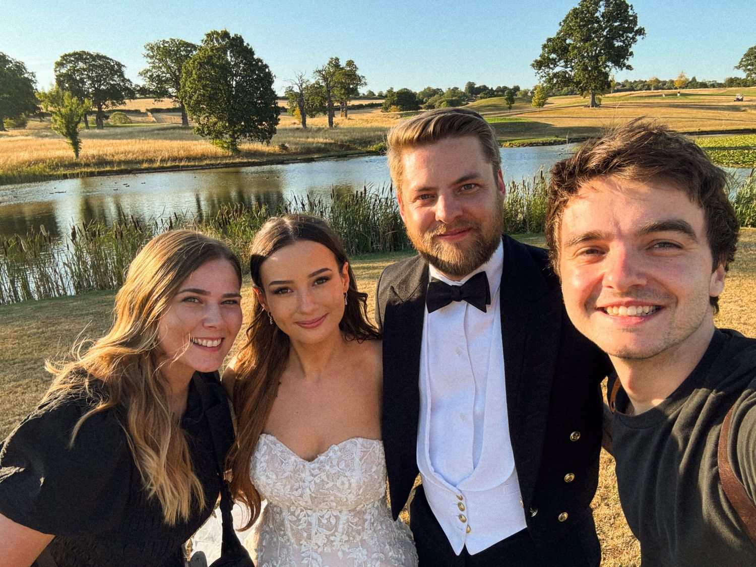 Two women and two men posing for a selfie outdoors at a wedding, with a scenic pond and trees in the background.