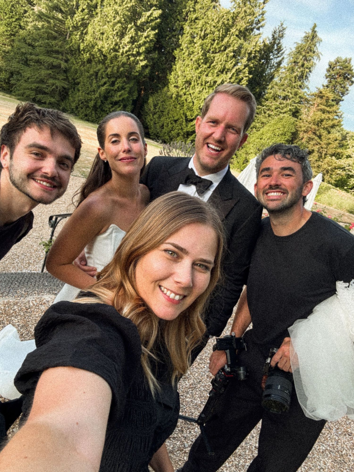 Group selfie of wedding couple in formal attire with three friends outdoors, surrounded by lush green trees.
