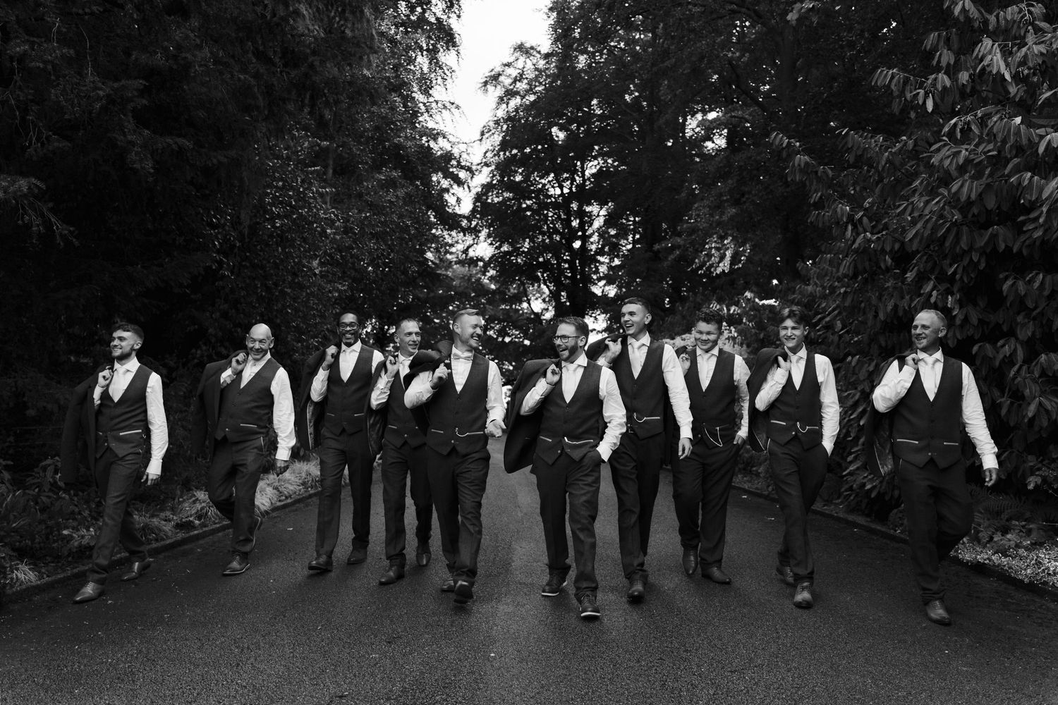Groomsmen in matching black vests walk together on a tree-lined path in black and white.