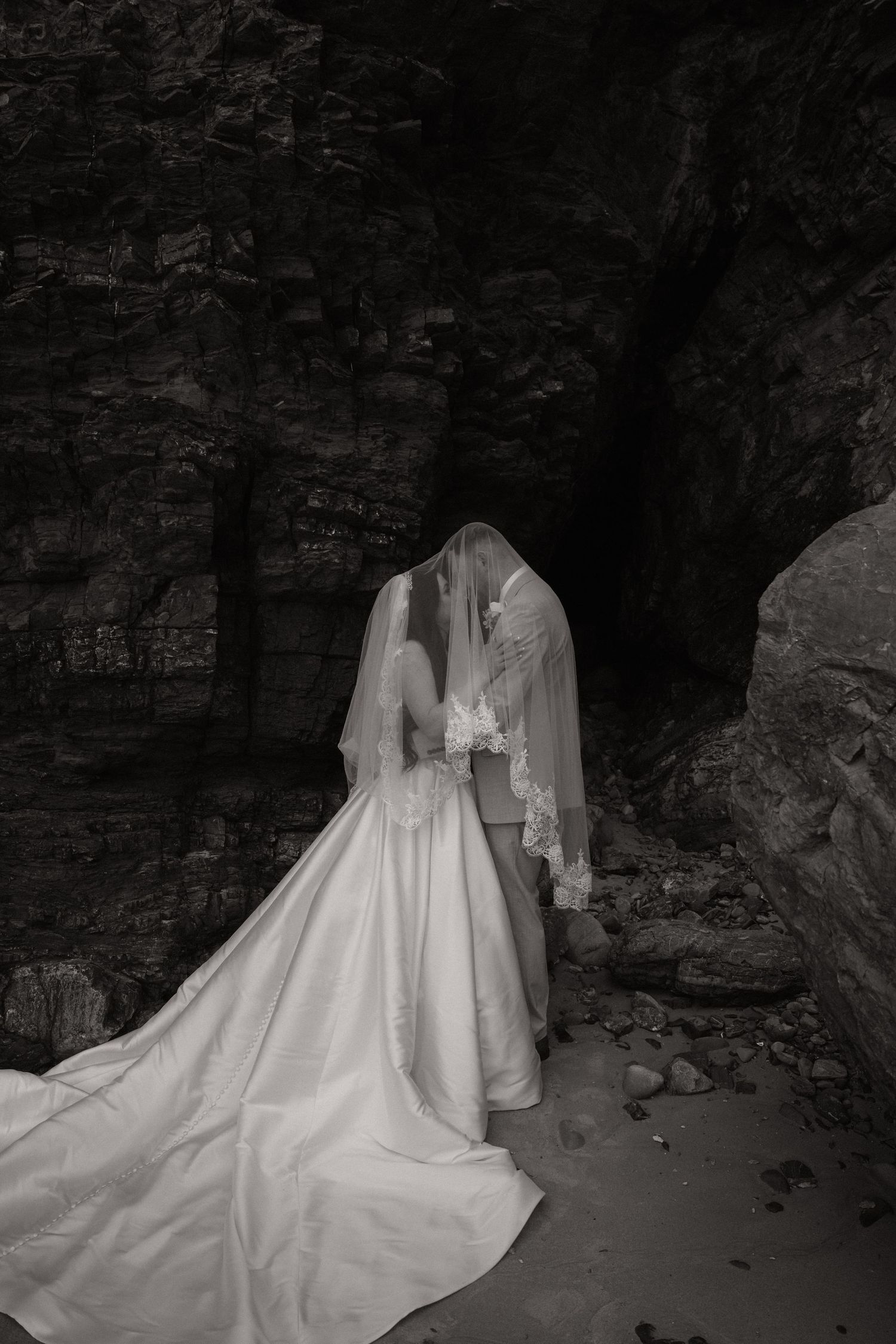 Black and white photo of bride and groom sharing a kiss under a rocky cave arch, bridal veil draped over both.