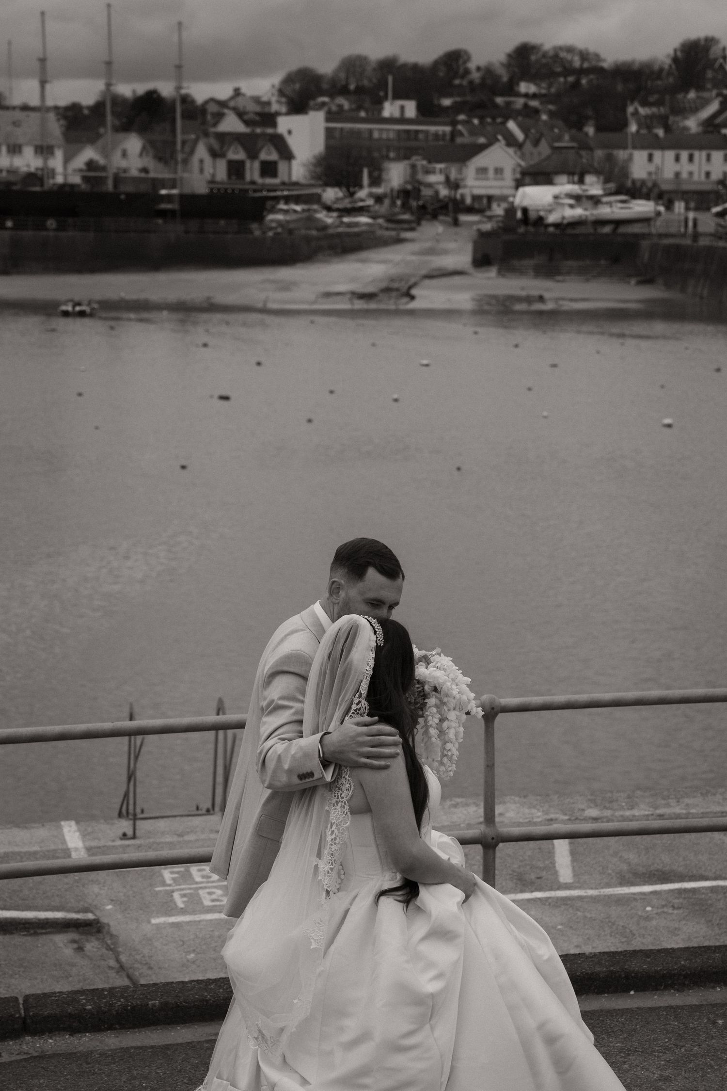 Black and white photo of a couple embracing on a waterfront railing, bride in flowing gown holding bouquet, harbor town backdrop.