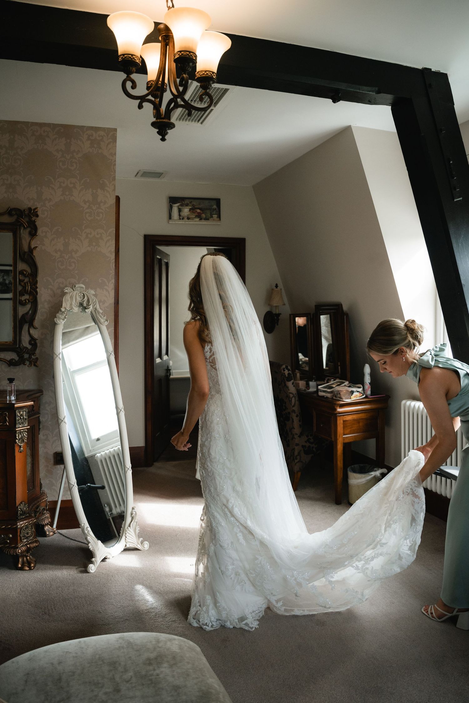 A wedding dress with a flowing lace train is displayed in a sunlit dressing room with vintage mirrors and decor.