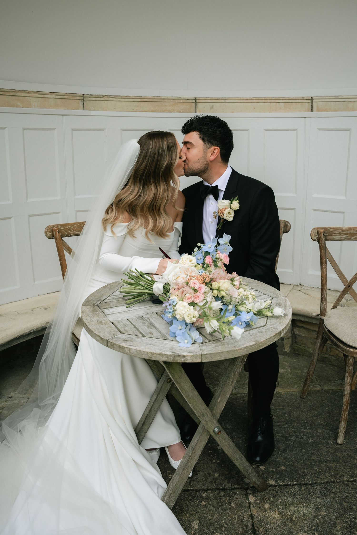 A romantic wedding scene with a couple sitting on rustic wooden chairs with delicate floral arrangements.