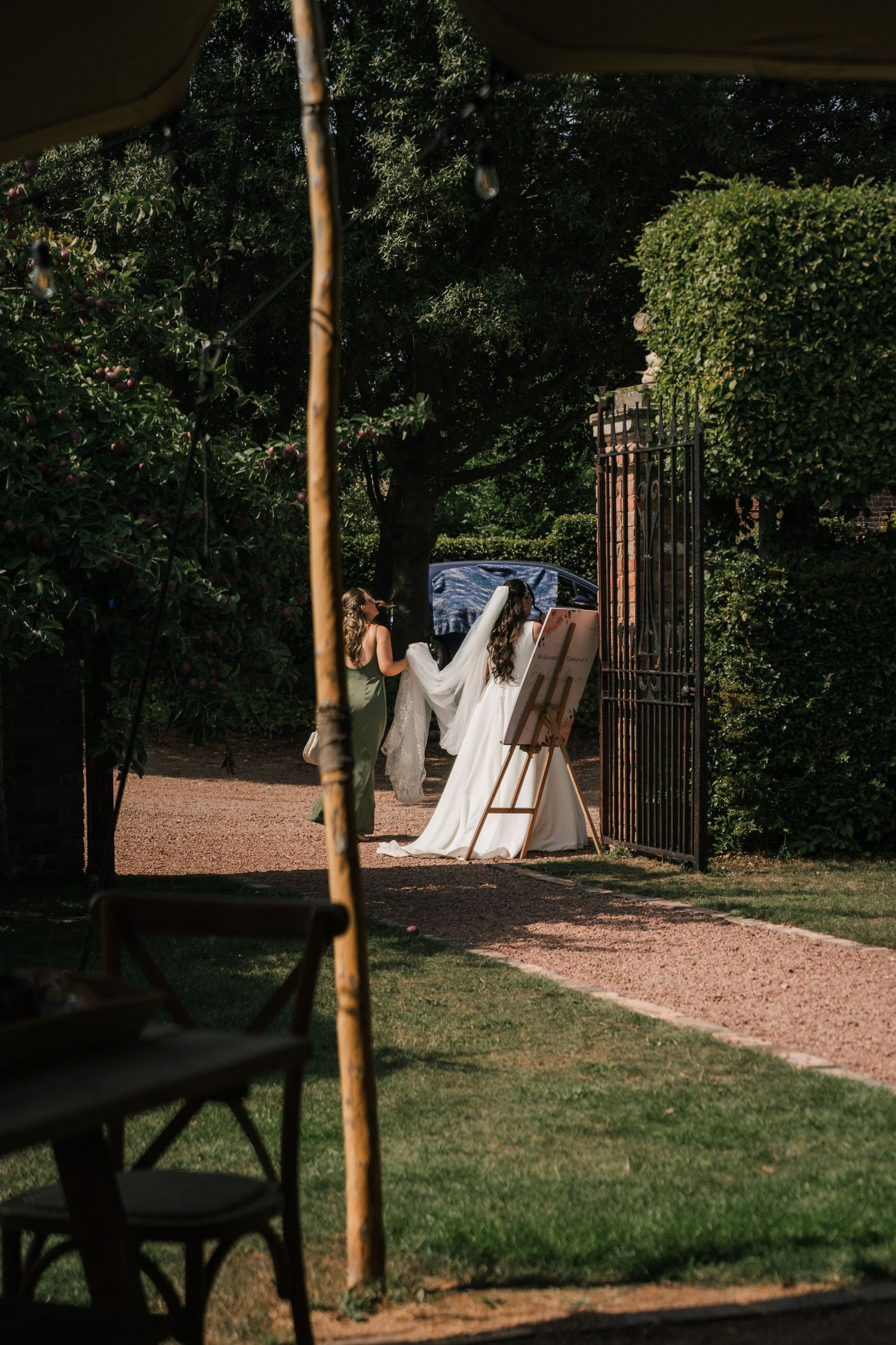White wedding dress hanging on a decorative iron gate along a garden path with trees and hedges in the background.