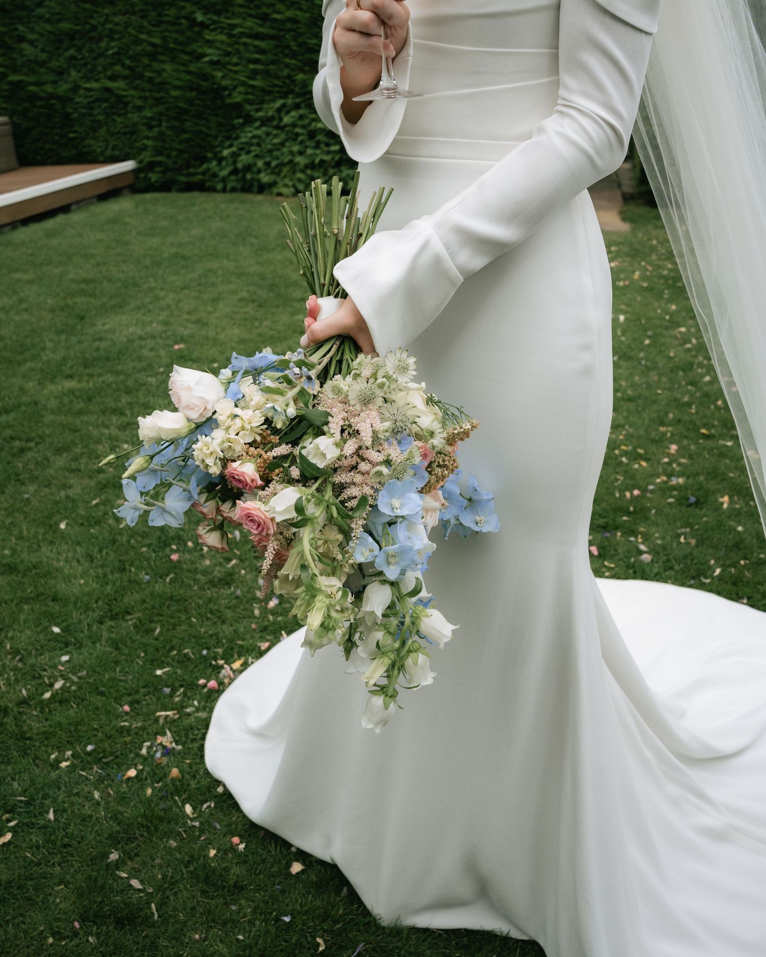 Elegant bride in white dress holding delicate bouquet with blue and pink flowers in garden setting.
