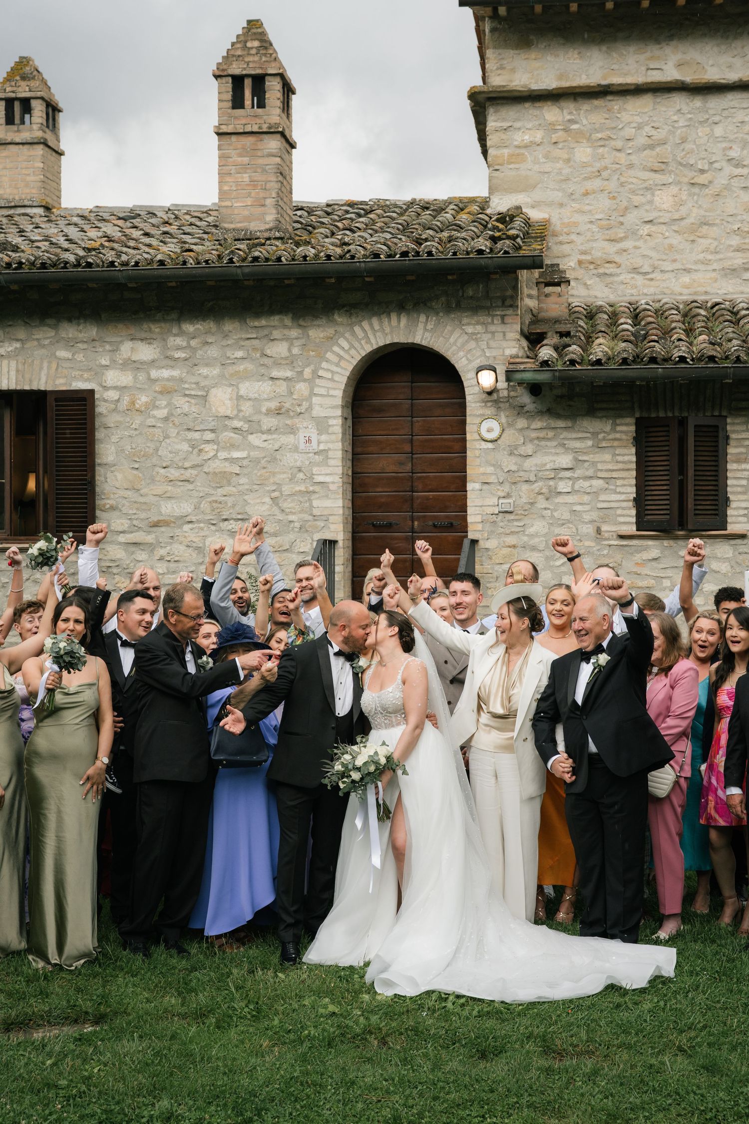 Large wedding party poses together on grass lawn in front of rustic stone building for group photo.