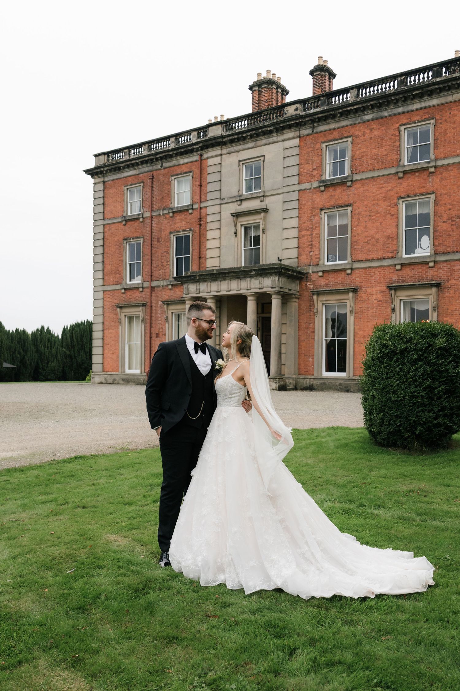 A couple in wedding attire poses in front of a grand historic brick mansion with manicured grounds.