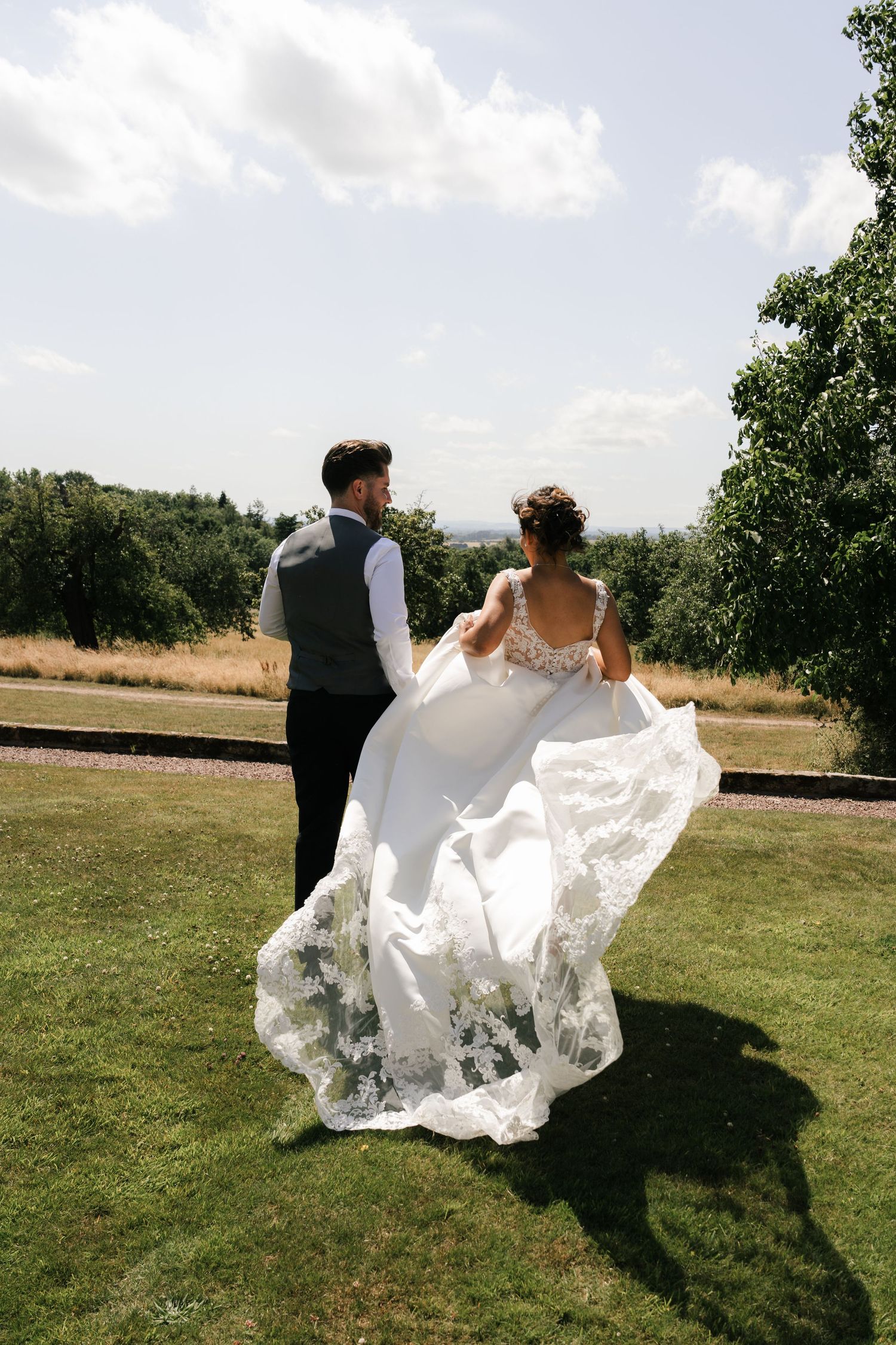 A wedding couple runs playfully through a grassy field as the bride's white dress flows in the wind.