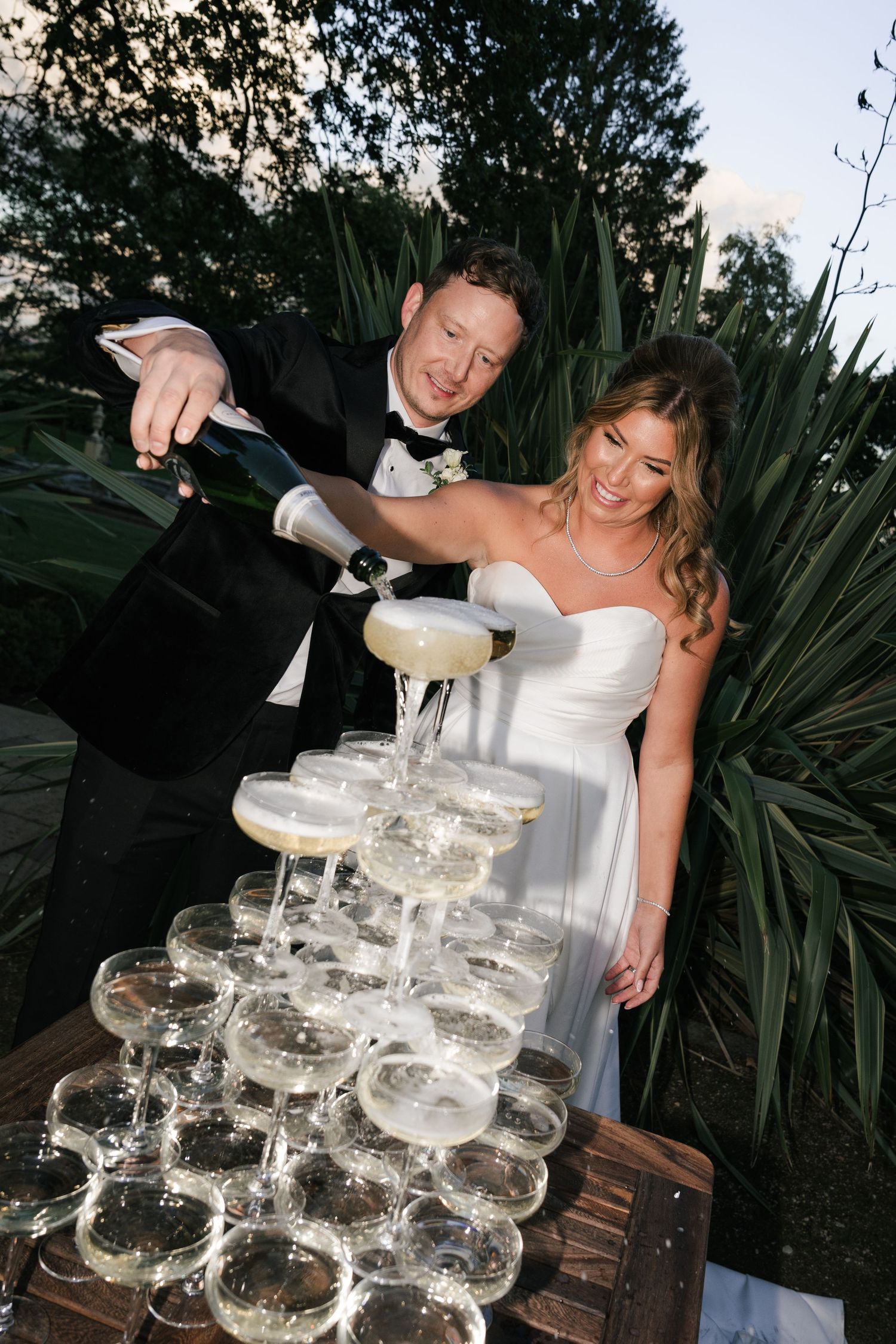 A champagne tower being carefully filled at an outdoor wedding celebration with glasses arranged in elegant tiers.