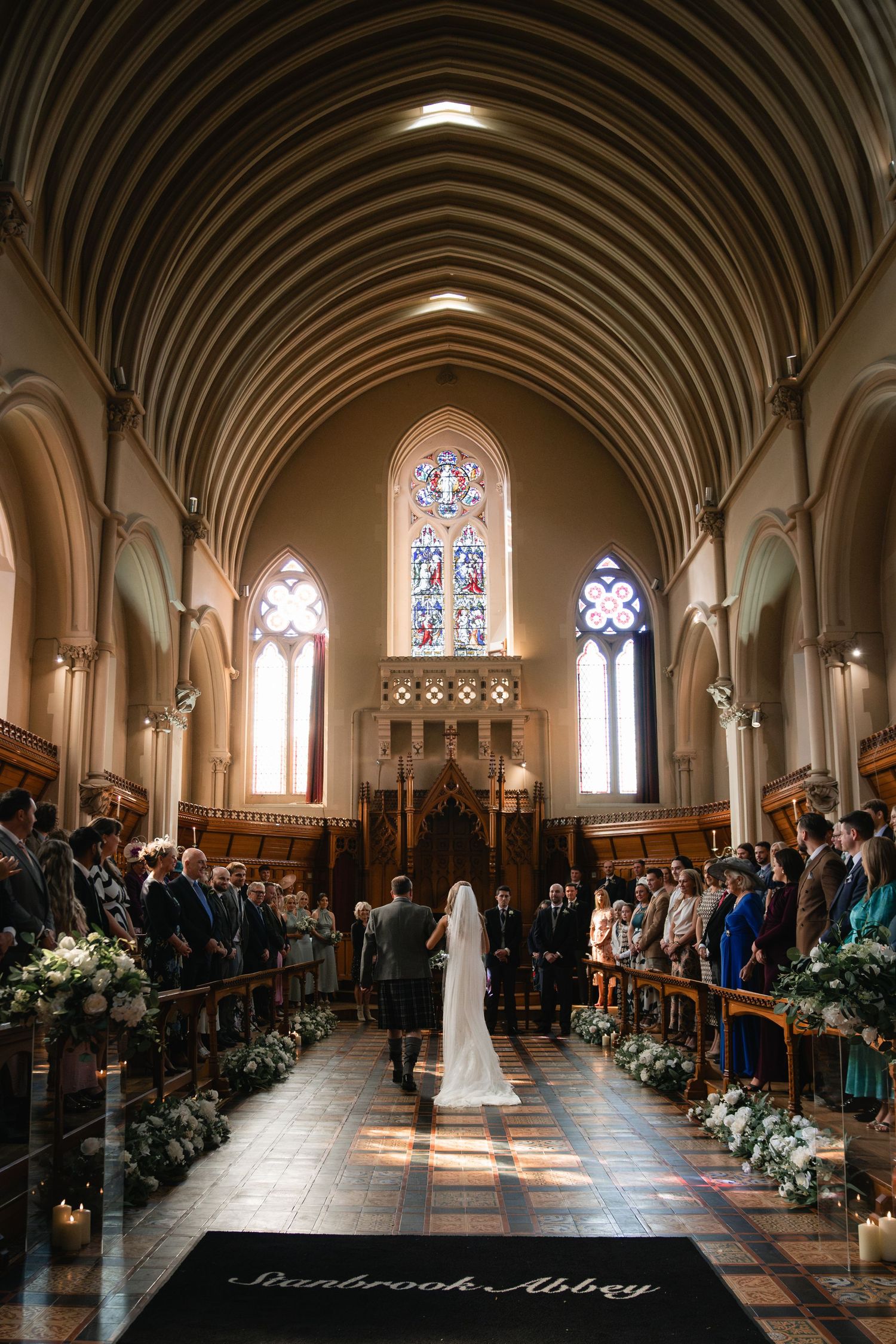 A wedding ceremony in a grand cathedral with stained glass windows, vaulted arches, and flower-lined aisle.