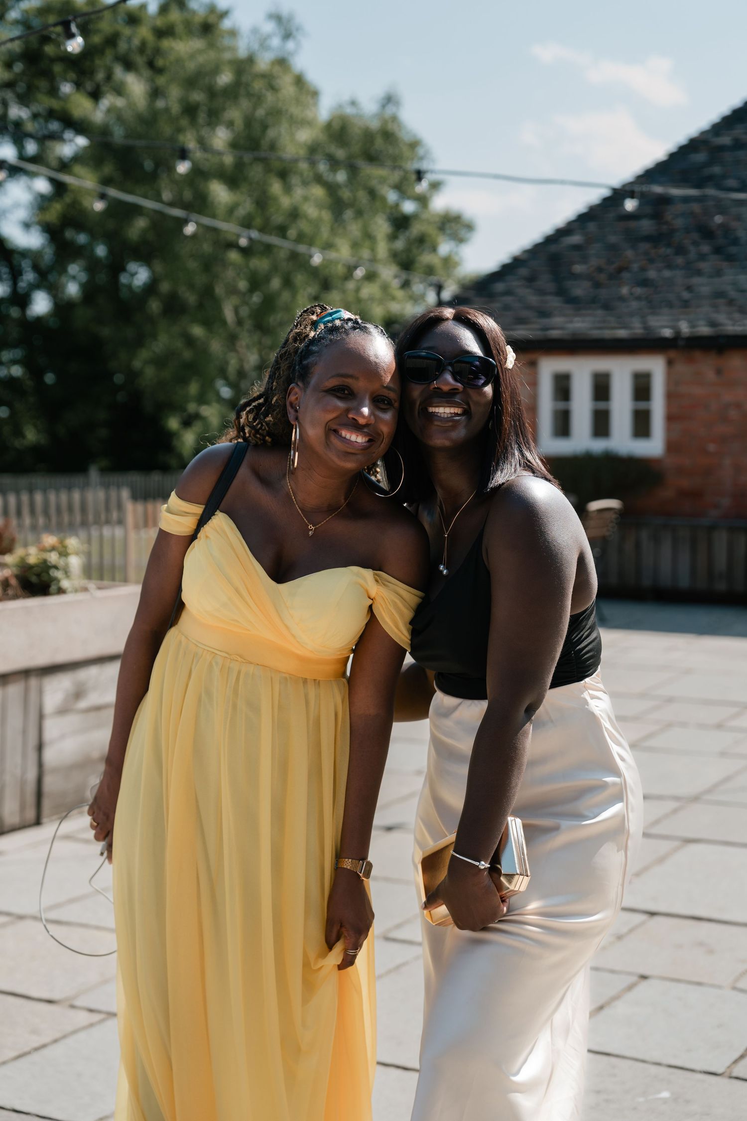Two friends share a joyful moment outdoors, one wearing a yellow dress and the other in black, against a brick building.