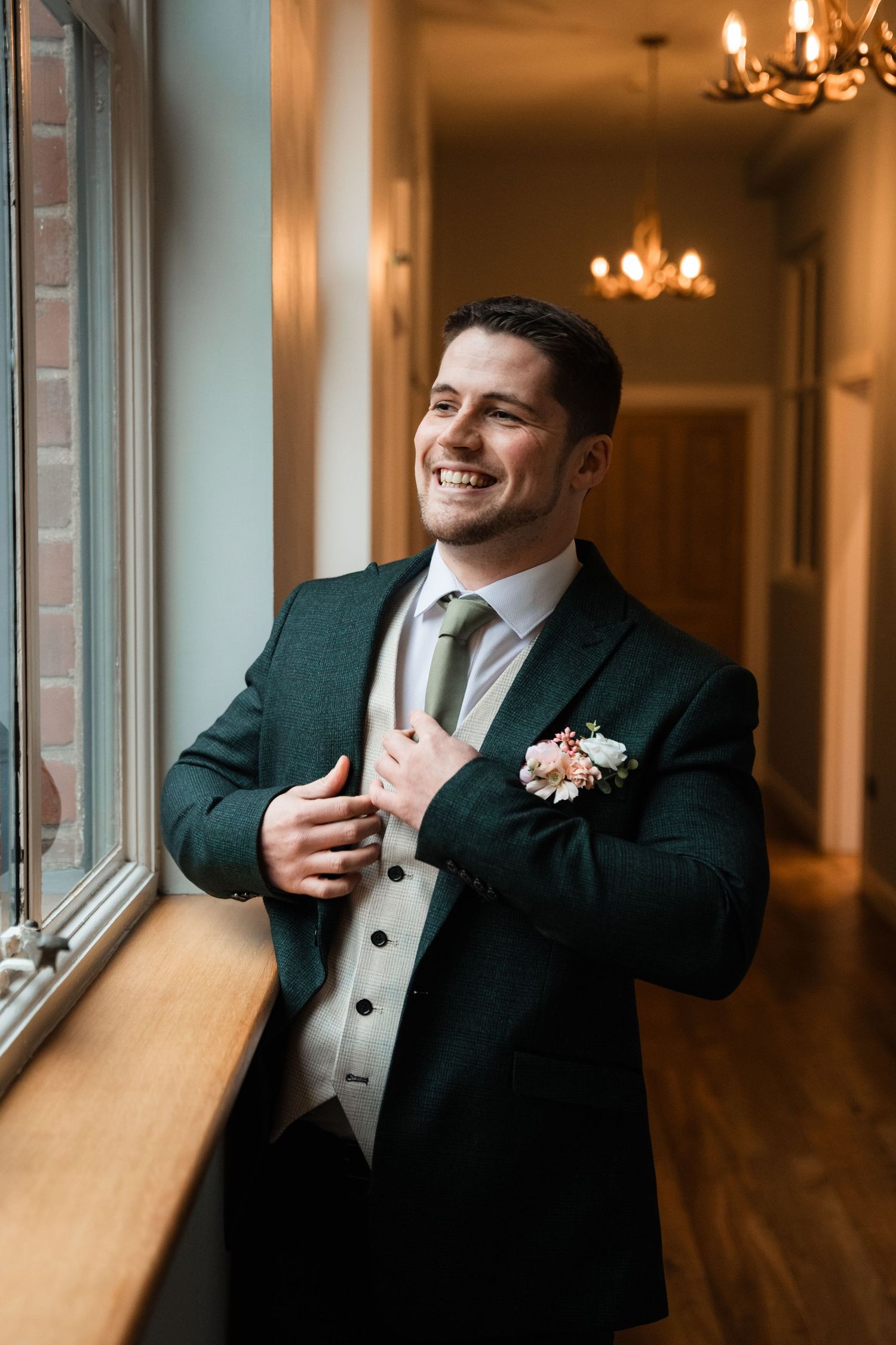 Groom in black tuxedo and white vest laughs while getting ready in elegant venue with chandeliers and windows.