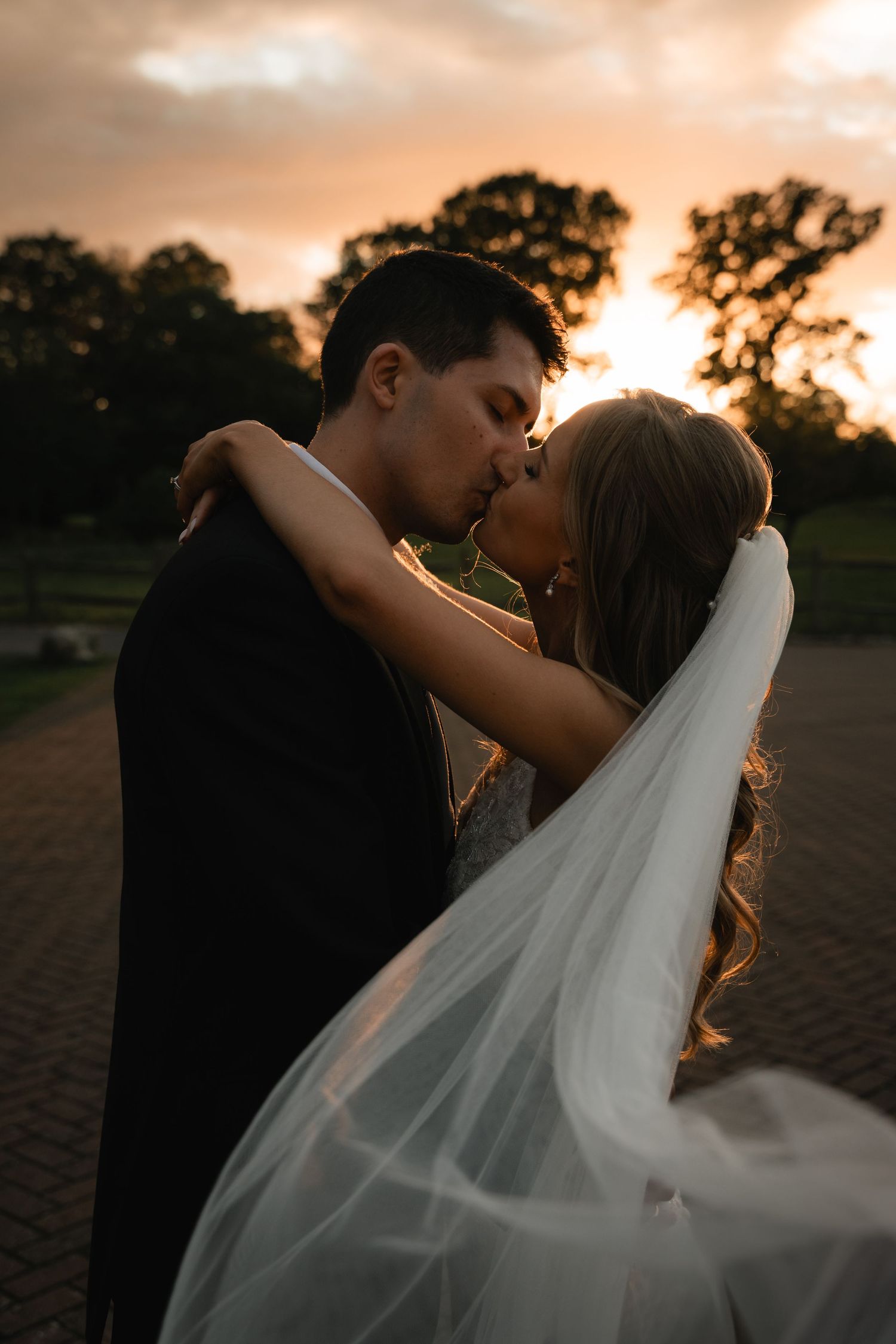 A romantic sunset silhouette of a bride and groom embracing with her flowing veil caught in the golden light.