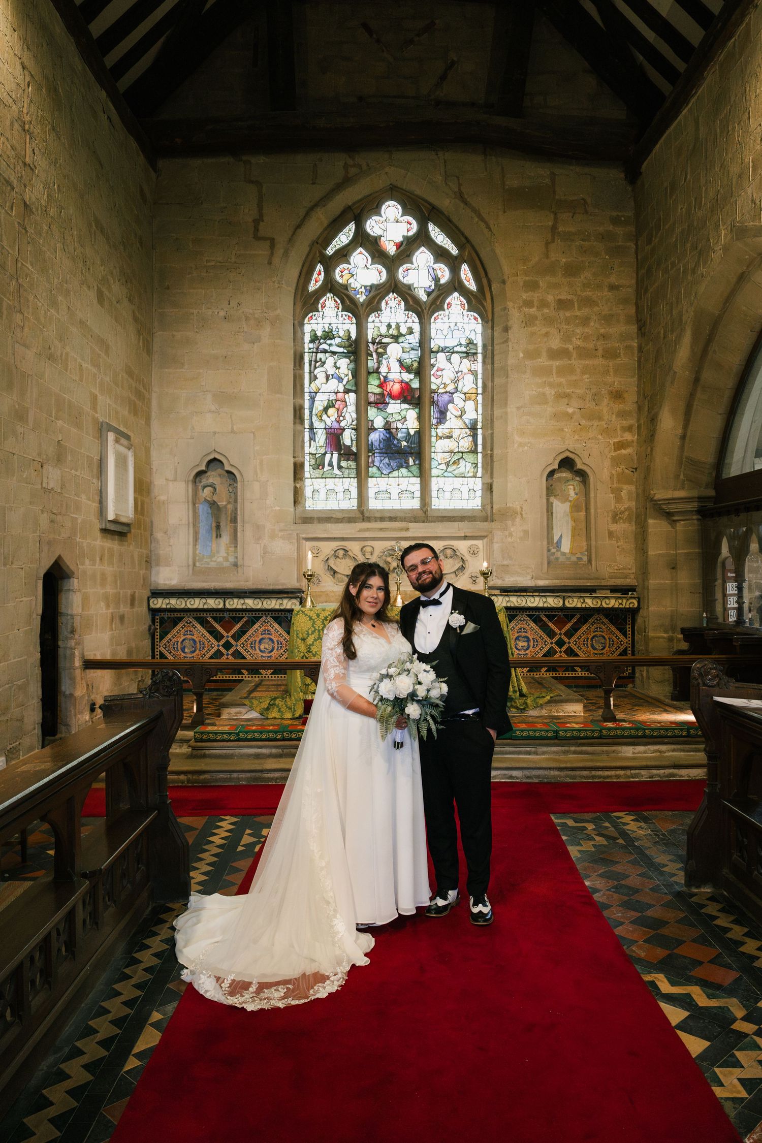 Newlyweds stand together in a historic church with stained glass windows and red carpeted aisle during wedding ceremony.