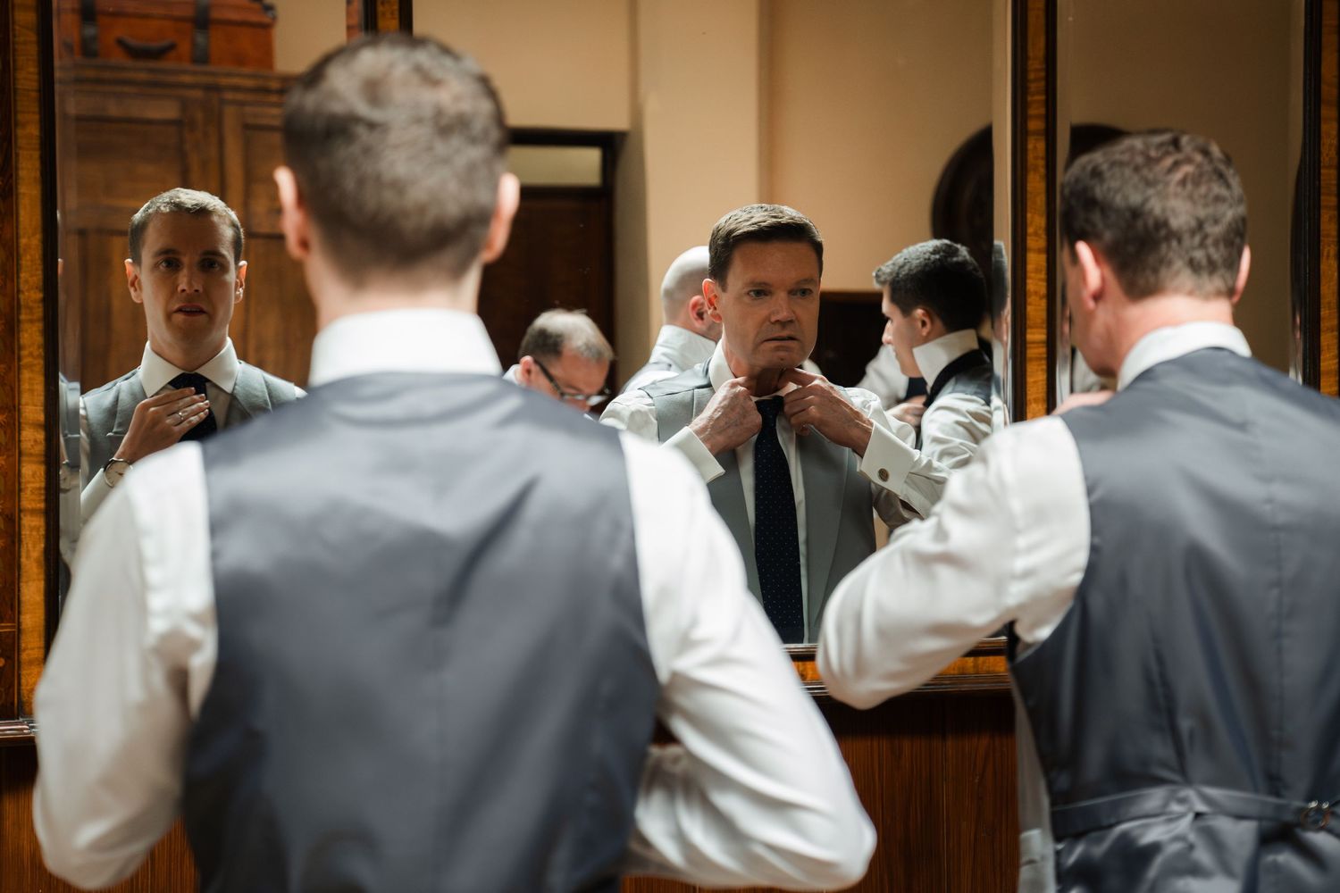 A group of groomsmen in suits and white shirts help each other get ready before a wedding ceremony.