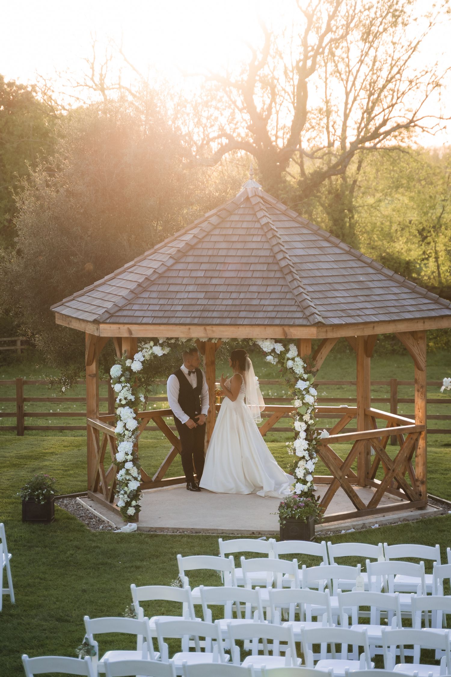 A rustic wooden gazebo decorated with white floral arrangements and chairs set up for an outdoor wedding ceremony at sunset.